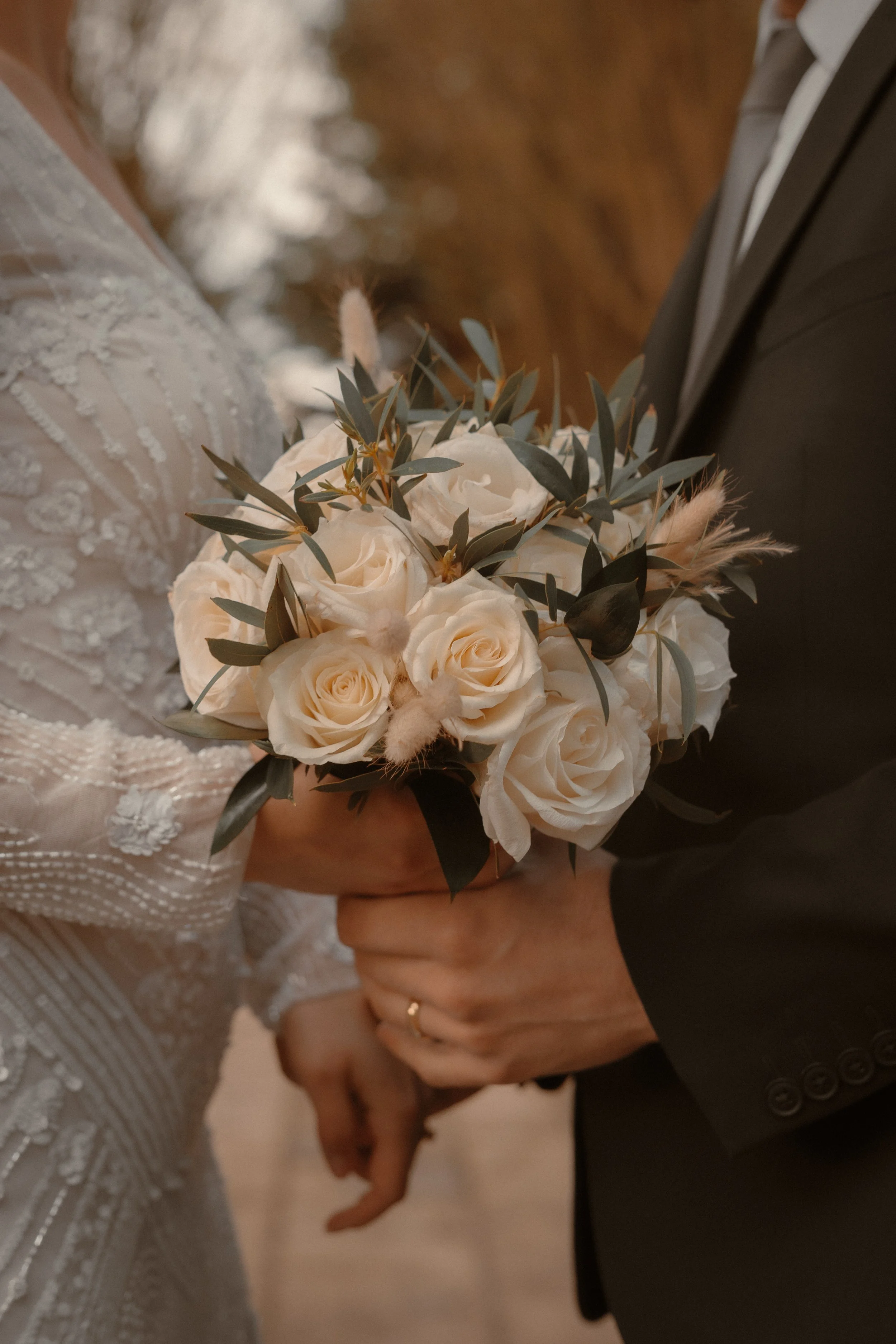 A bride and groom holding a bouquet of white roses with greenery and decorative dried elements, during their wedding.