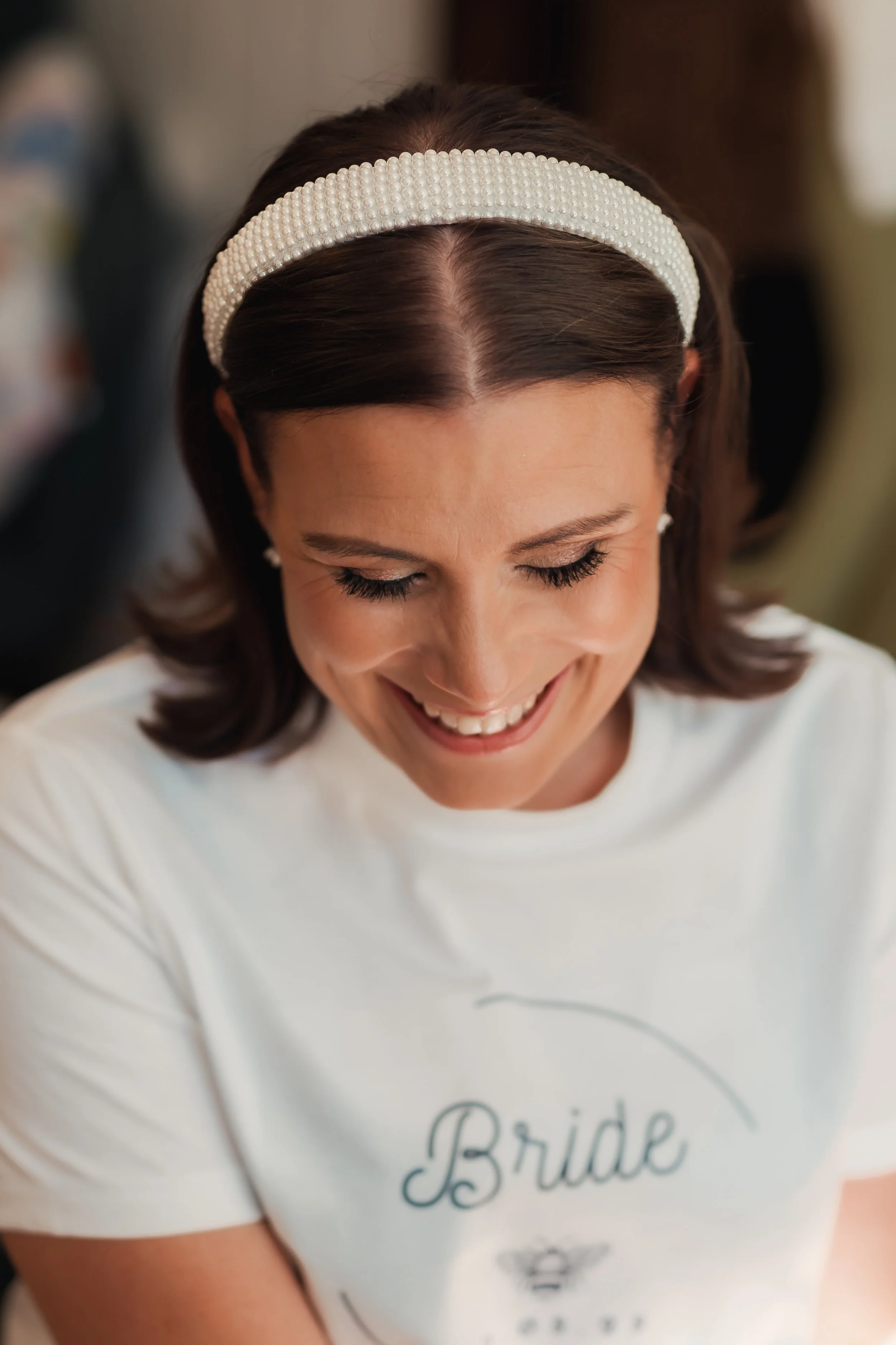 A smiling woman wearing a pearl headband, white earrings, and a white T-shirt with the word 'Bride' on it, looking down with eyes closed.