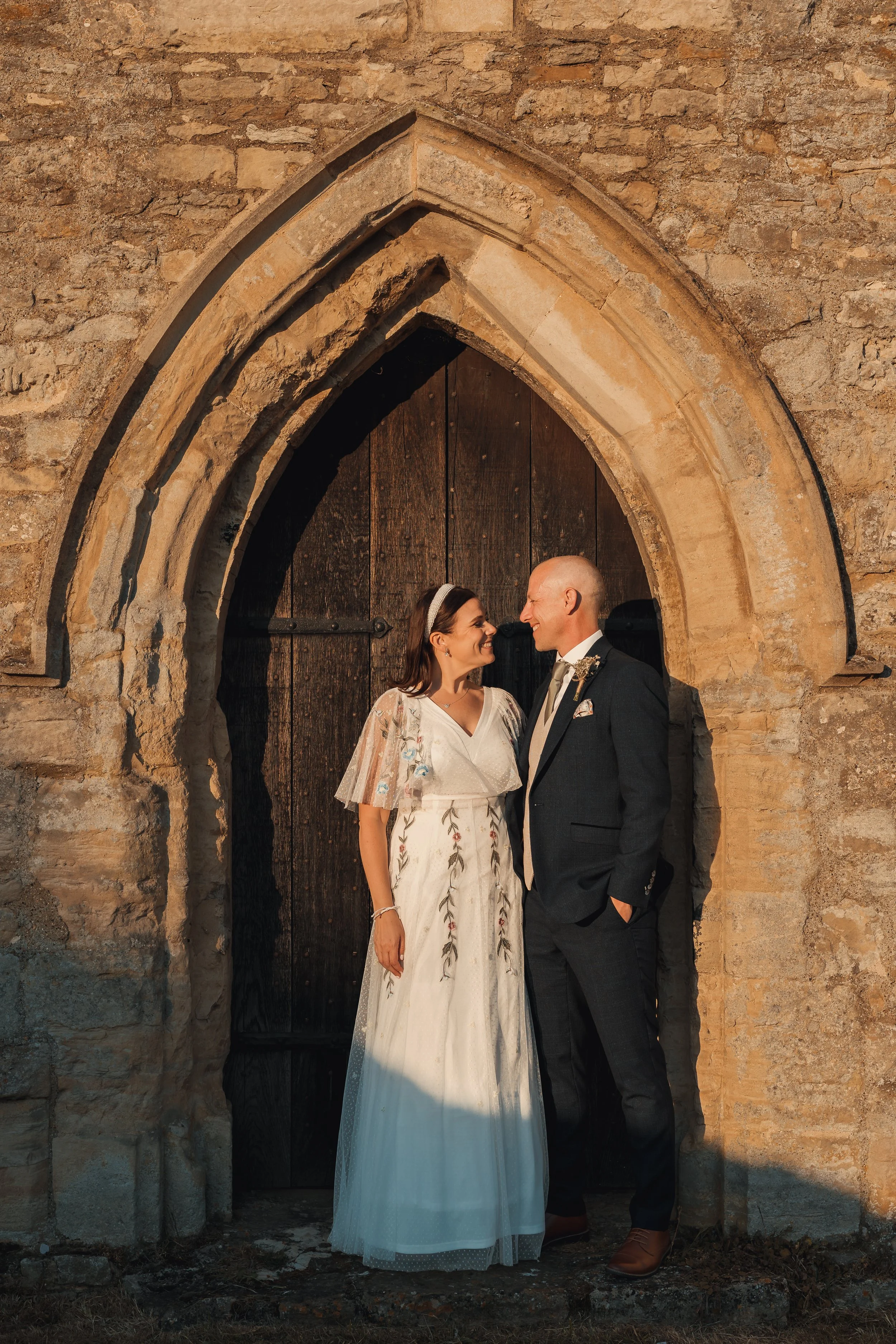 A newlywed couple wearing wedding attire standing close together in front of a dark wooden church door framed by an arched stone doorway, warmly lit by sunset.