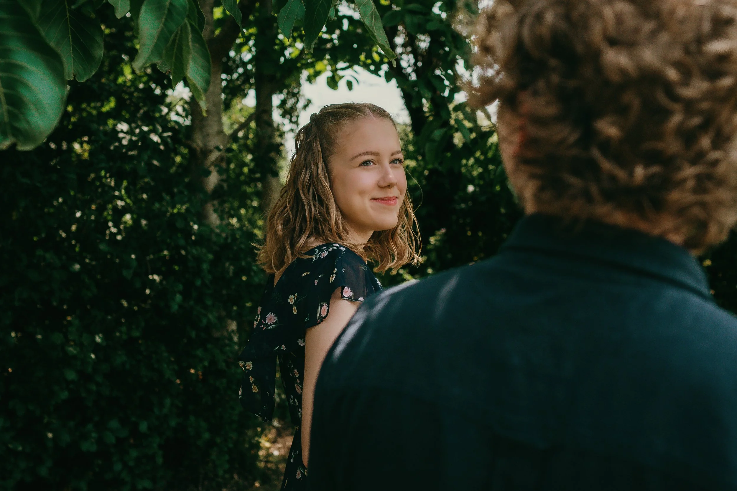 A young woman with shoulder-length wavy hair, smiling and looking at an older person with curly hair, outdoors among lush green trees.