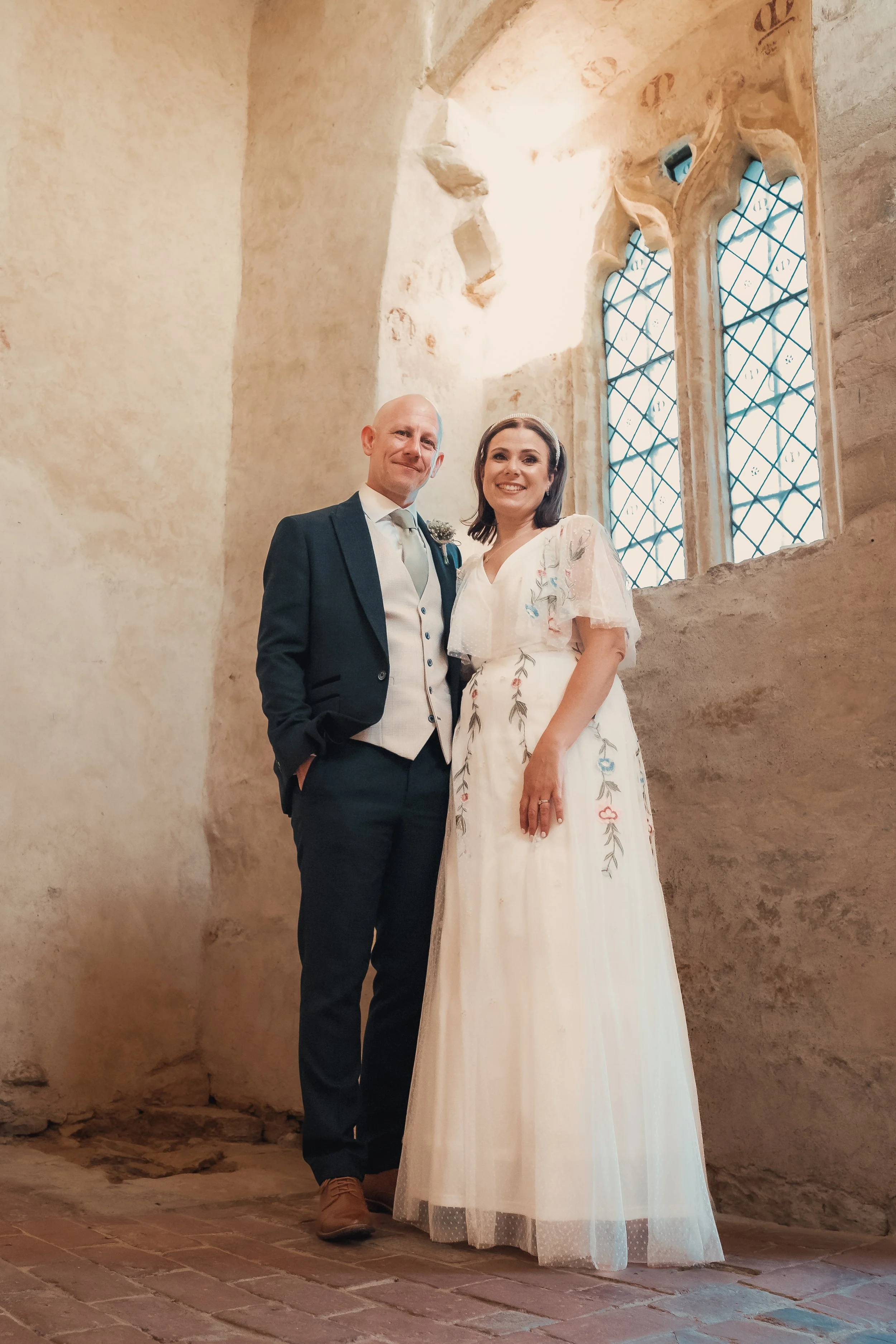 A bride and groom standing inside a historic stone church with stained glass windows during their wedding.