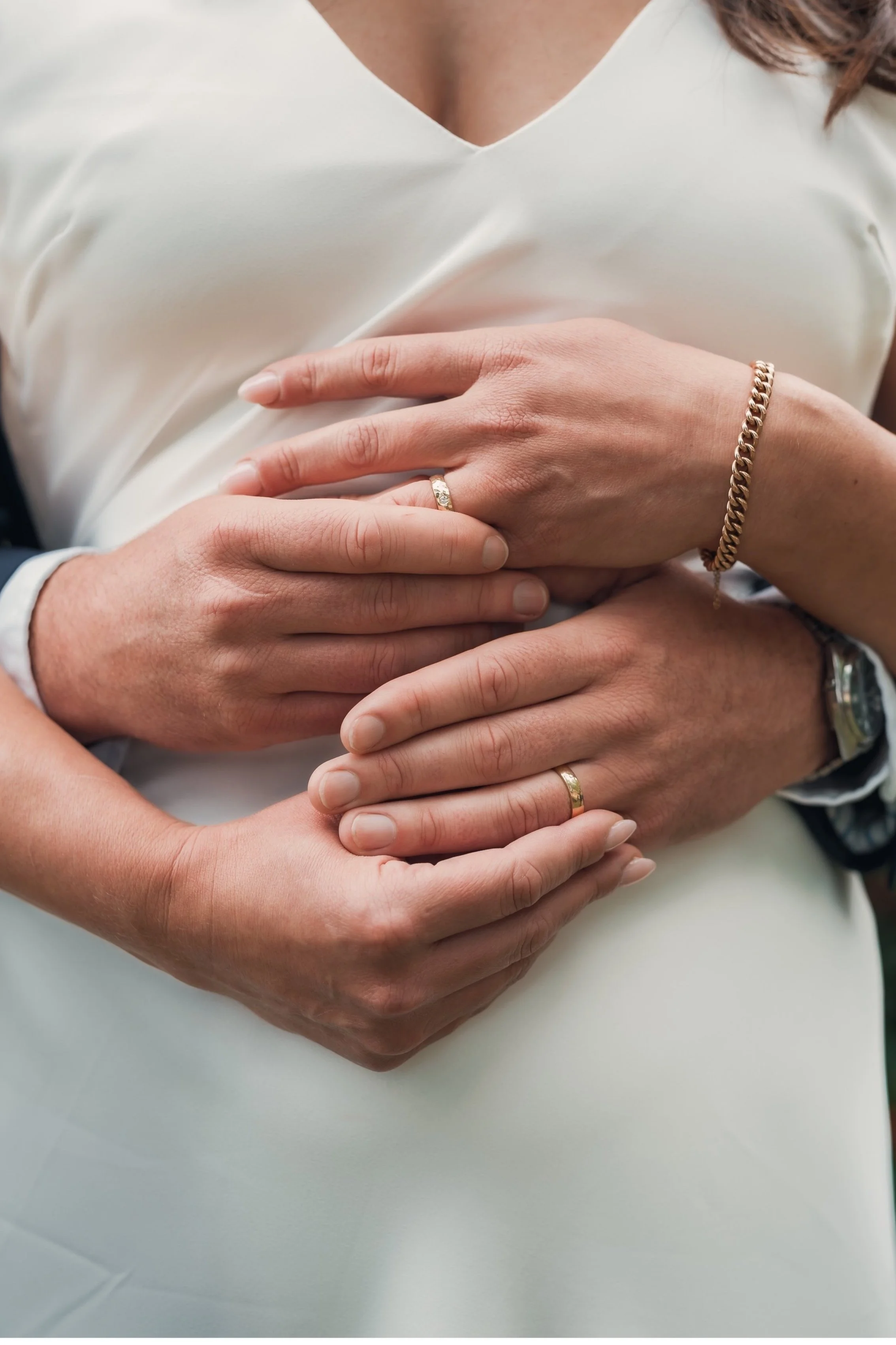 Close-up of a couple’s hands, showing wedding rings and jewelry, embracing on a woman in a white dress.