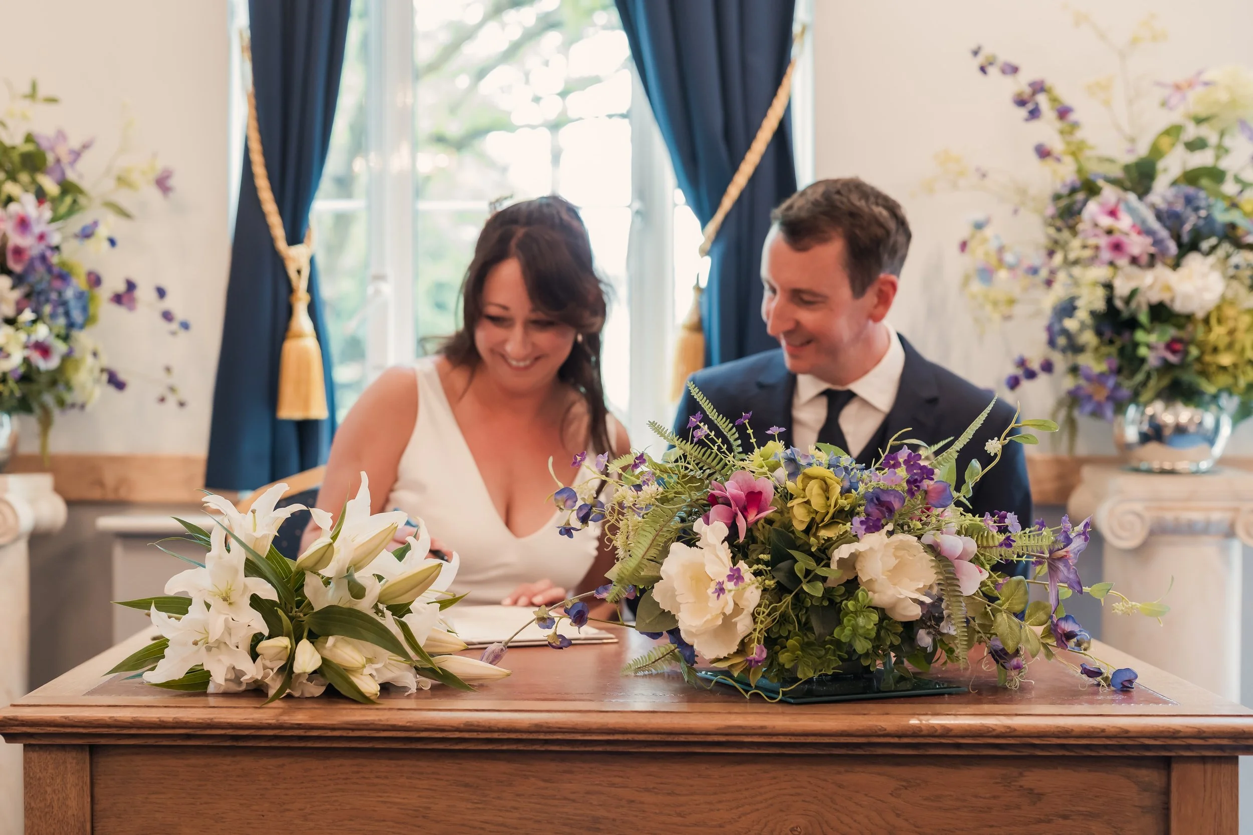 A bride and groom signing their wedding registry or legal documents at a decorated table, with bouquets of white and colorful flowers in front of them, inside a room with large windows and elegant blue curtains.