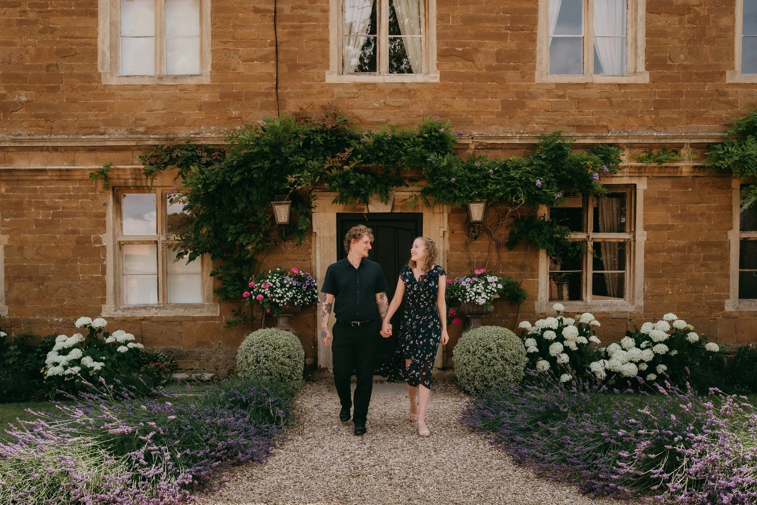 A couple holding hands and walking on a gravel path in front of a brick house with lush greenery and flowers.