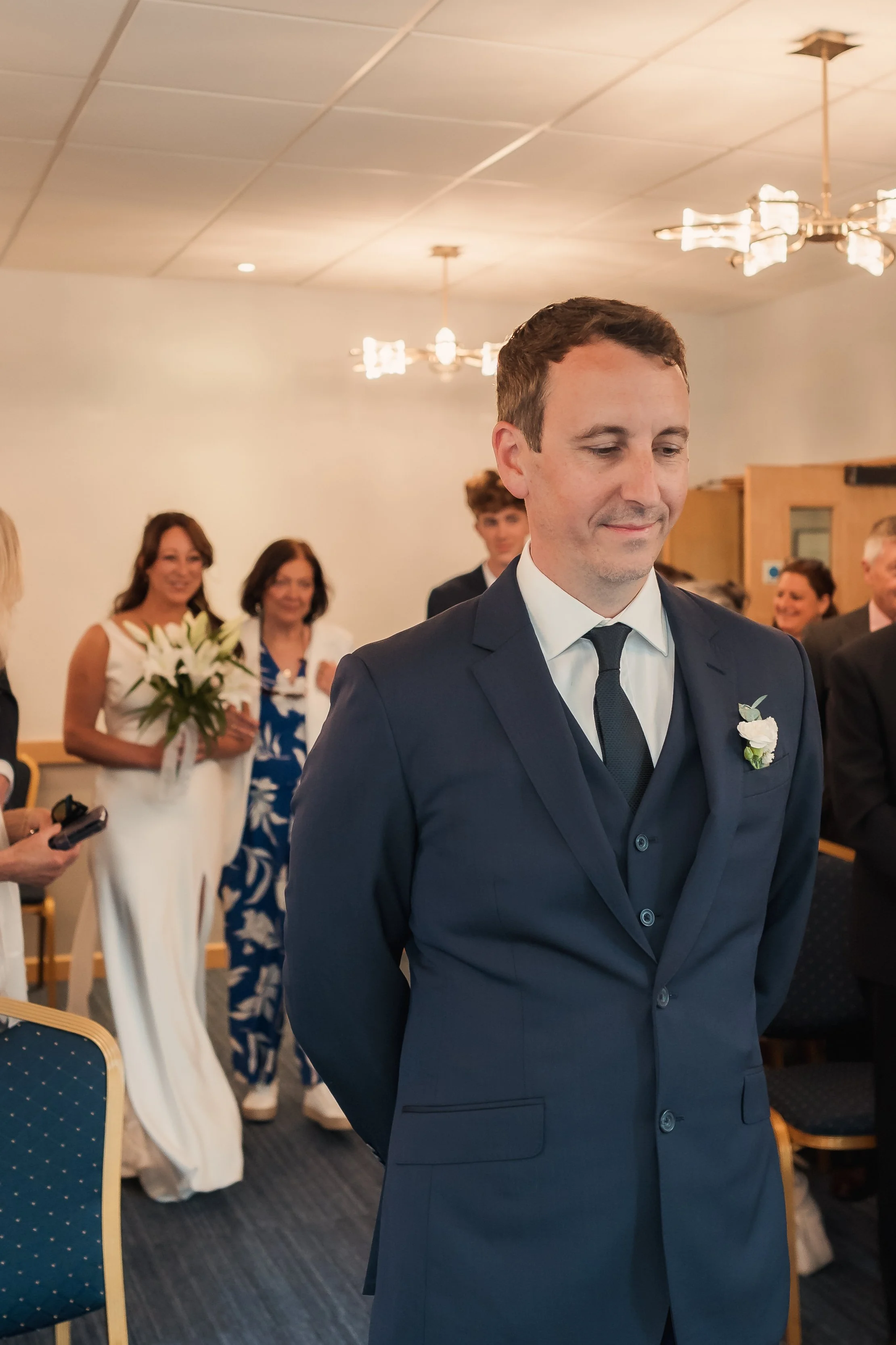 A groom in a navy suit with a white boutonniere stands with his head bowed during a wedding ceremony, with guests in formal attire standing behind him in a room with chandeliers and wooden accents.