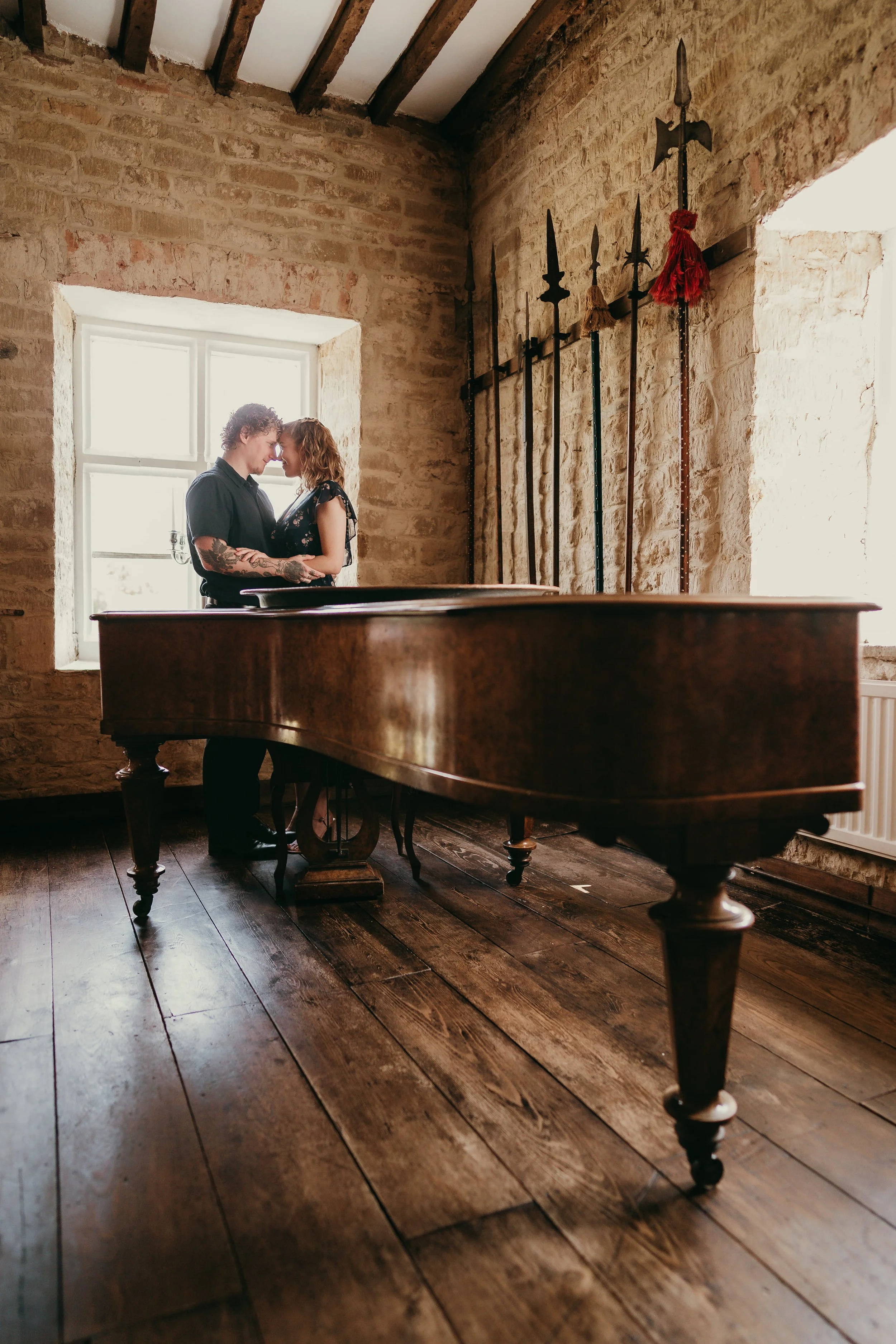 A couple standing close together inside a rustic room, with a large wooden table in the foreground and a band of weapons hanging on the brick wall behind them, illuminated by natural light from a window.