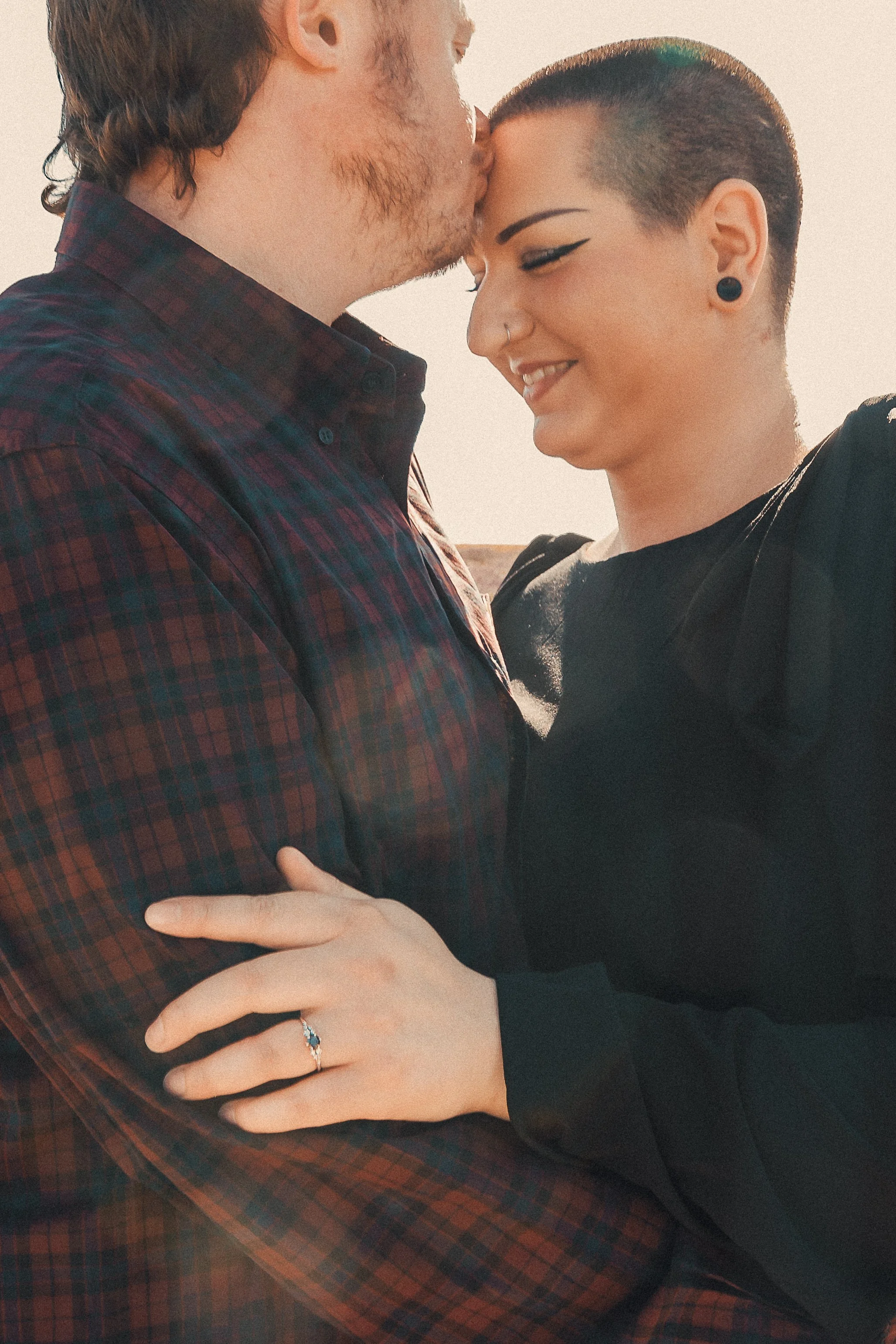 A man and woman embrace closely, with the man kissing the woman's forehead. The woman is smiling with a ring on her finger, and they are holding each other fondly outdoors in warm sunlight.