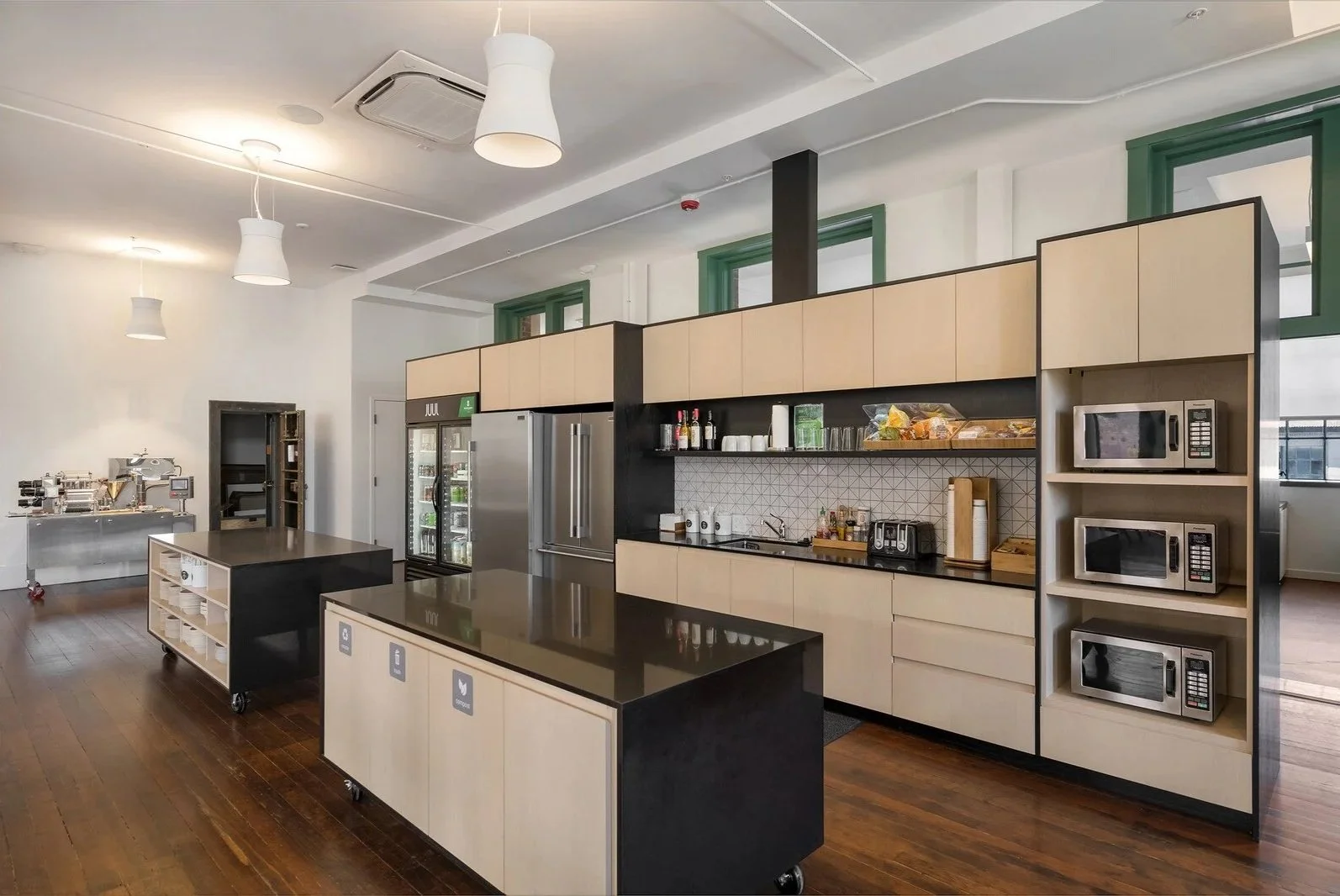 Union Iron Works Headquarters communal kitchen space in San Francisco with dark wood floors, white walls, modern black countertops, white cabinets, painted green trim, food, drinks, and appliances by Orton Development Inc.