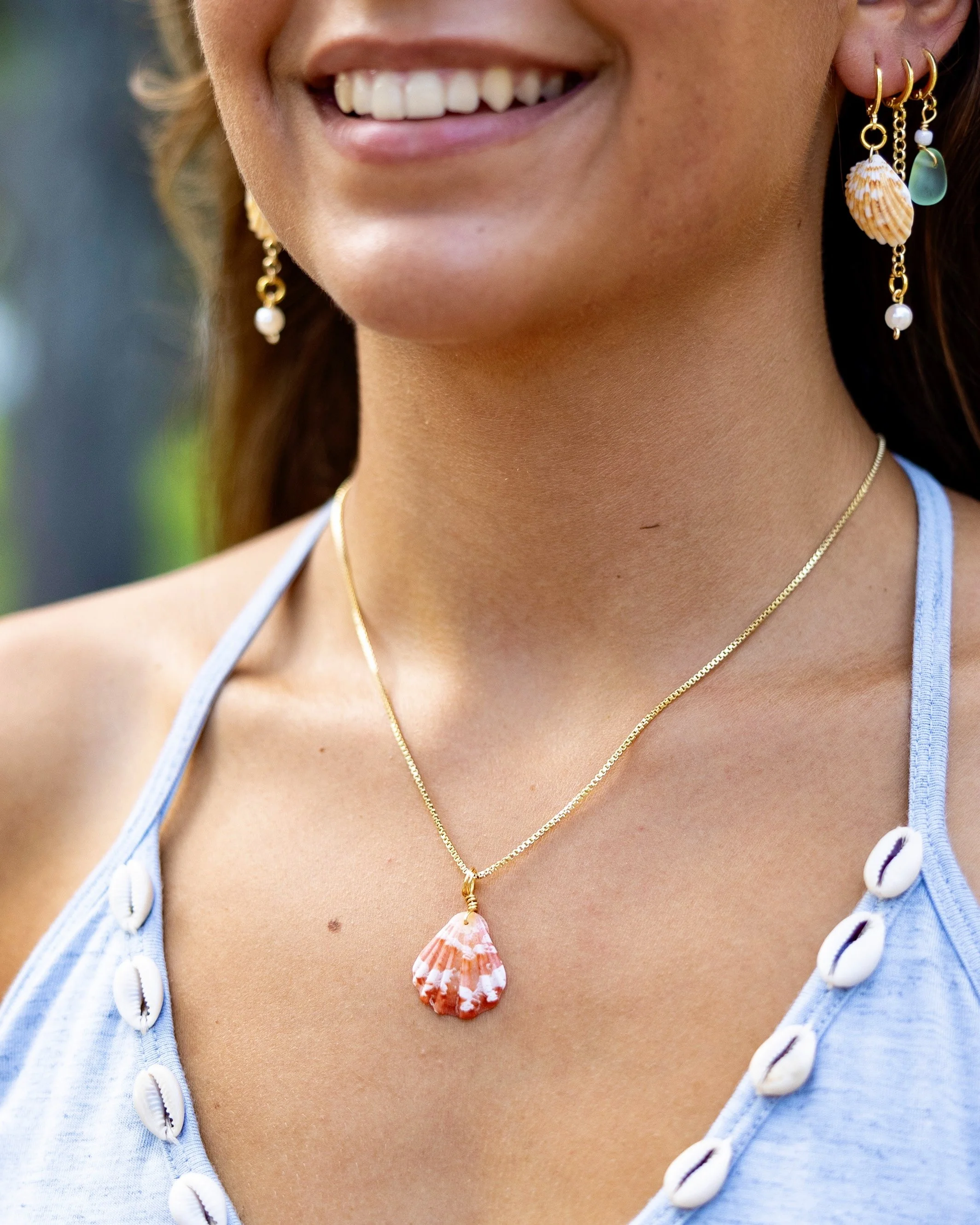 Close-up of a woman's face and necklace with a seashell pendant, wearing earrings with seashell and pearl charms, smiling, dressed in a blue top with shell decorations.
