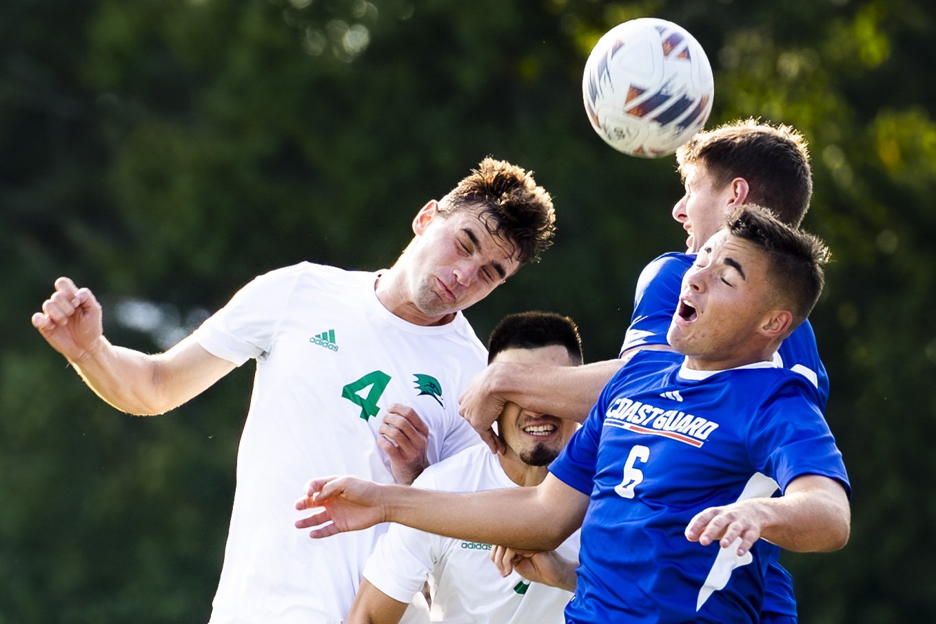 Four young men playing soccer outdoors, three wearing white jerseys and one in a blue jersey, competing for the ball in the air on a grassy field.