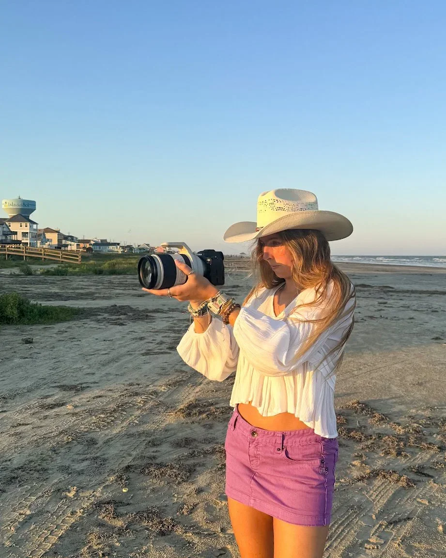A woman wearing a white cowboy hat, a white long-sleeve blouse, and a pink skirt is standing on a beach, looking at her camera as the sun sets.
