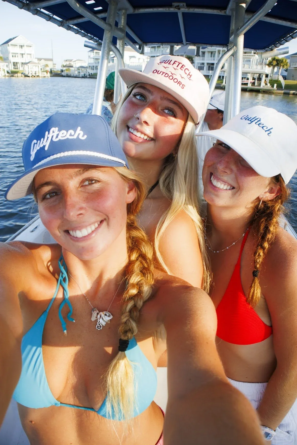 Three women taking a selfie on a boat, wearing caps with 'Gulftech' branding, smiling, with water and residential houses in the background.