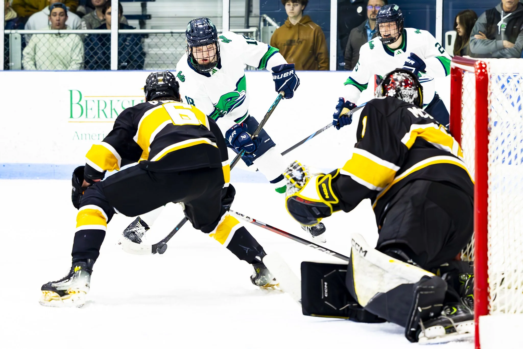 Hockey players in action during a game, with one player attempting to score while the goalie and defenders try to block the shot, on an ice rink.