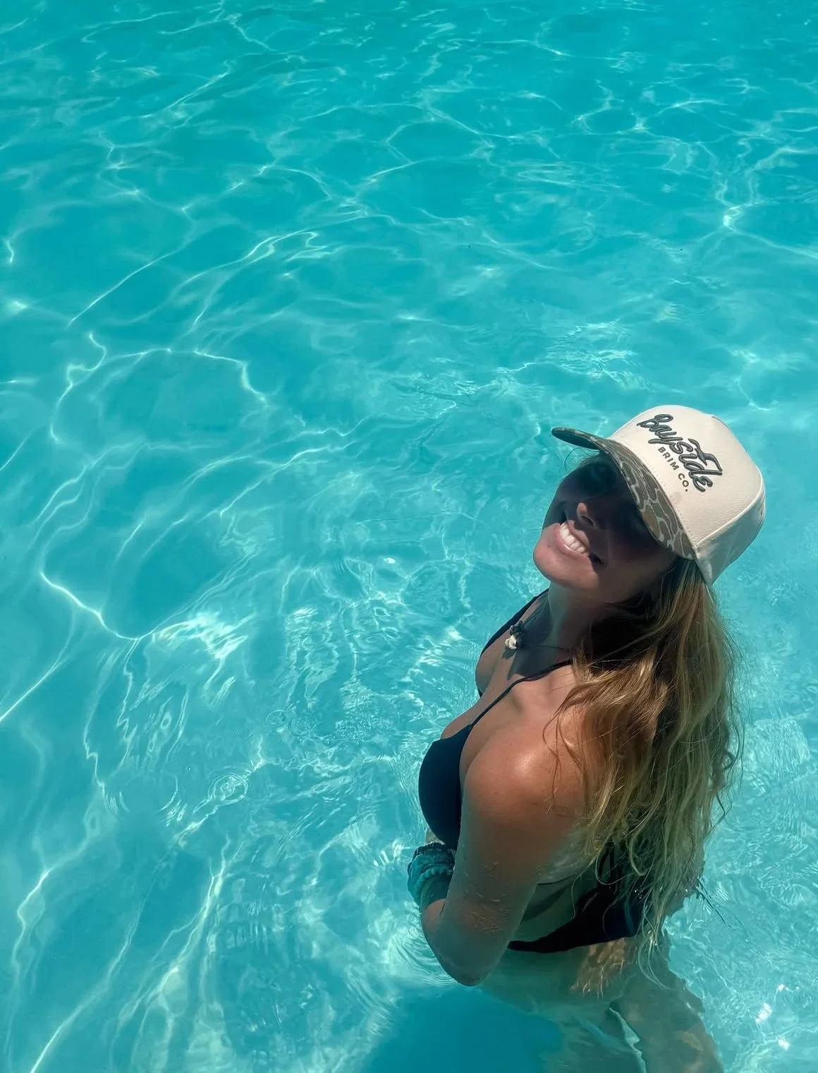 A woman in a swimming pool, wearing a black swimsuit, a white cap, and sunglasses, smiling with her teeth visible.