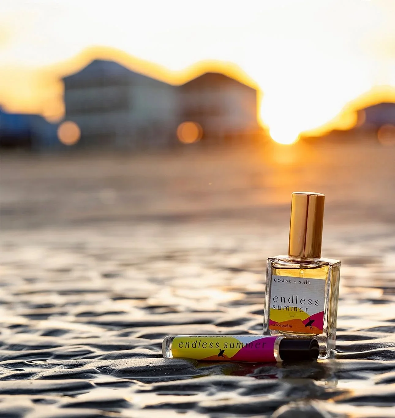 A bottle of perfume and a lip balm with the label 'endless summer' placed on wet textured ground with blurred houses in the background during sunset.