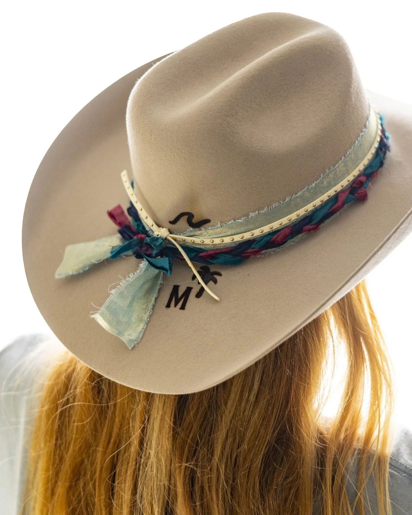 A beige cowboy hat with a decorative band and ribbon, worn by a person with long red hair.