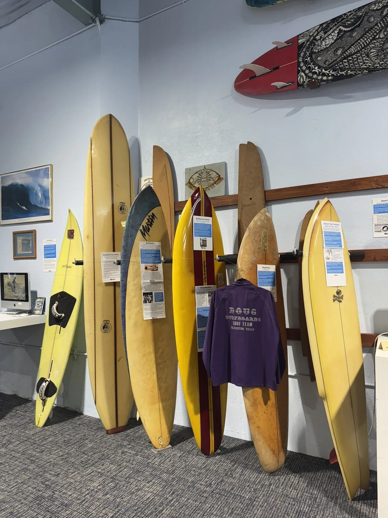 Collection of vintage surfboards and a purple surf team jacket displayed on a wall in a surf museum.
