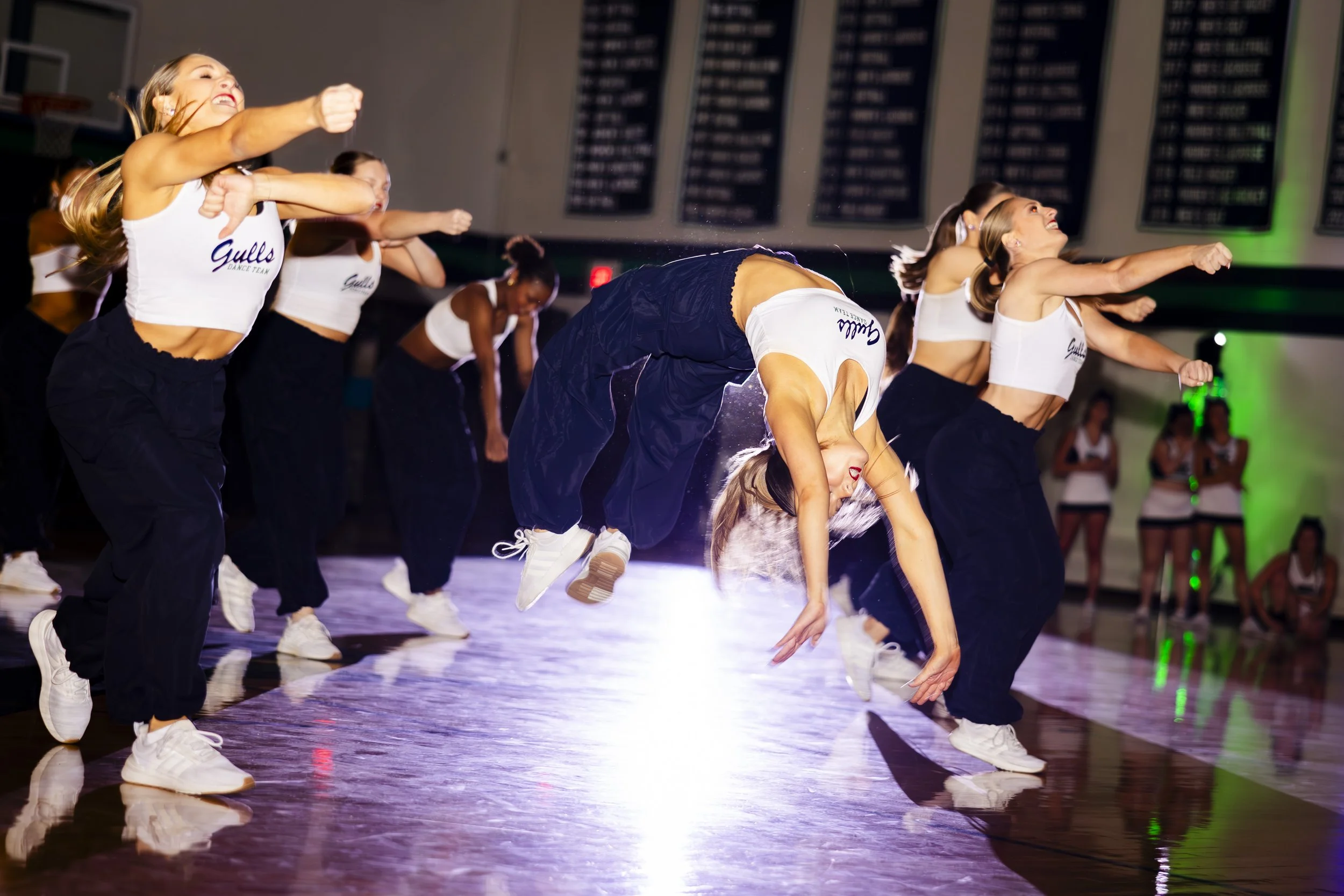 Cheerleaders performing a handstand flip on the gym floor during a dance practice or competition, wearing matching white crop tops and black pants.