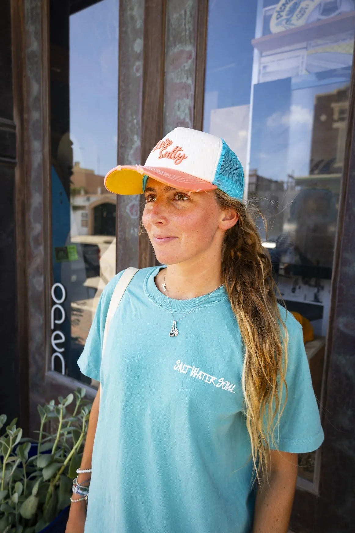 A young woman with long wavy hair wearing a trucker hat and a turquoise t-shirt with white text standing outside a business with a glass door, reflected buildings, and a blue sky.