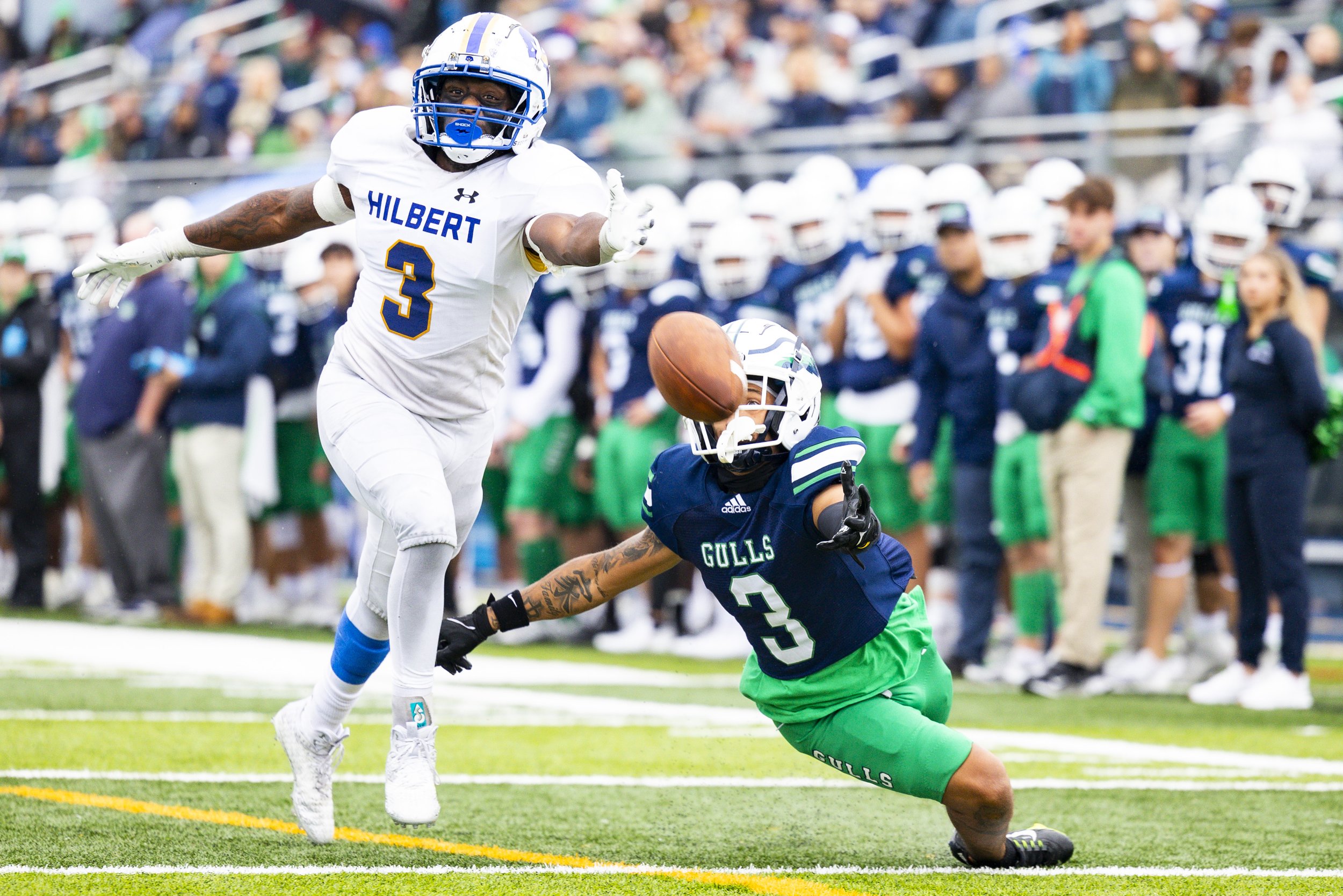 A football game with a player in a white jersey and a player in a navy jersey reaching for the football on the ground.