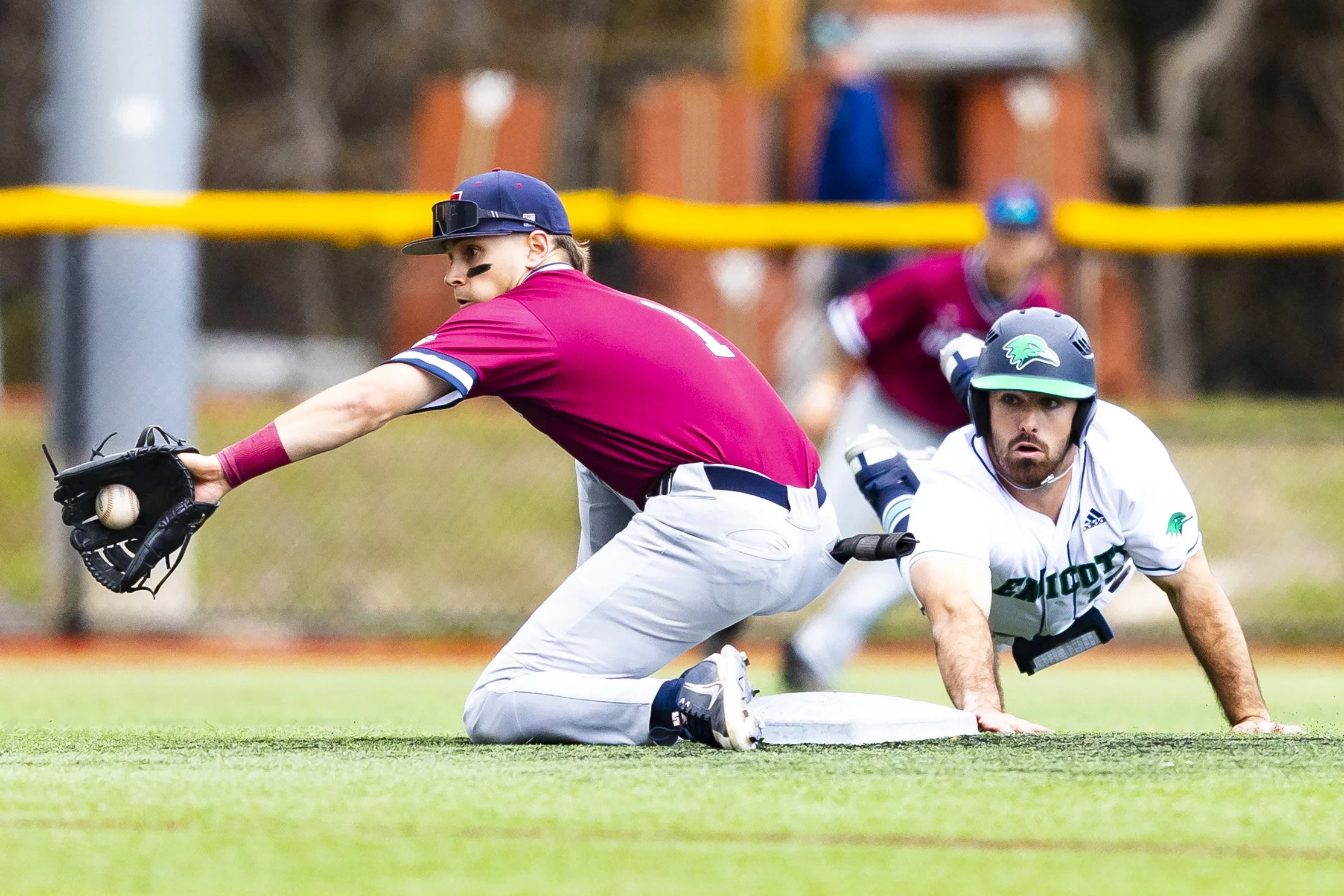 A baseball player wearing a pink jersey and gray pants dives to second base, reaching for the ball in his glove, while the baseman, in a white jersey with a green helmet, is on his hands and knees looking towards him. A player in a maroon jersey is v