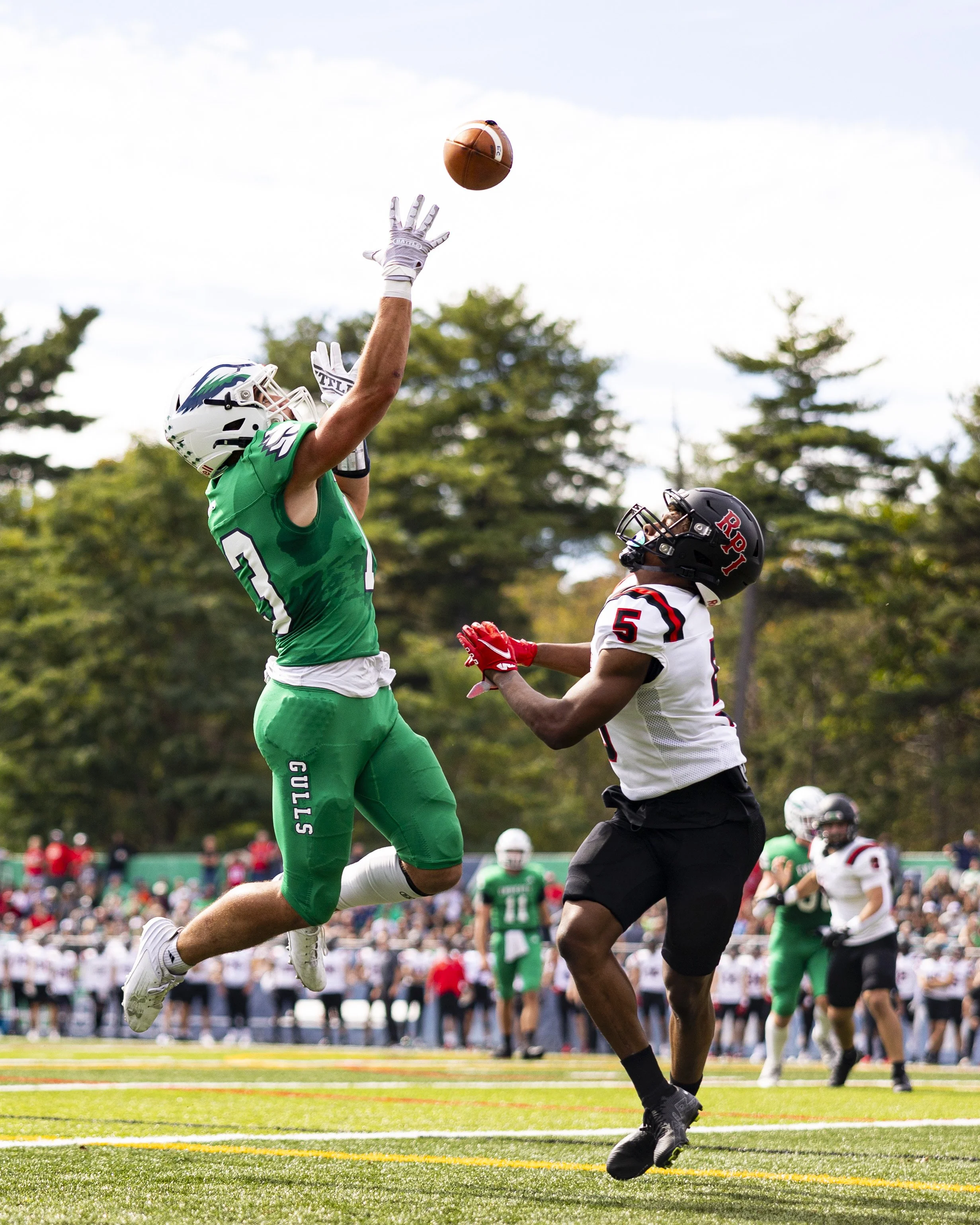 Football player in a green uniform jumping to catch the ball with an opposing player in a white uniform and black helmet attempting to block him during a game.
