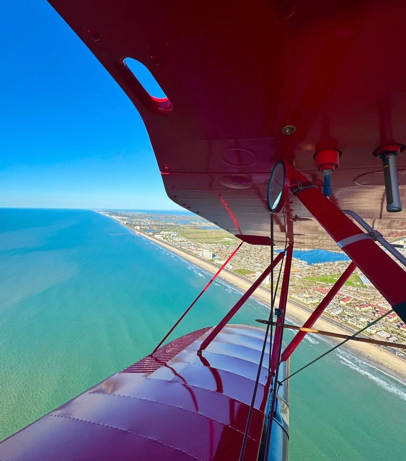 View from an airplane flying over the coastline, showing the ocean, beach, and land with buildings.