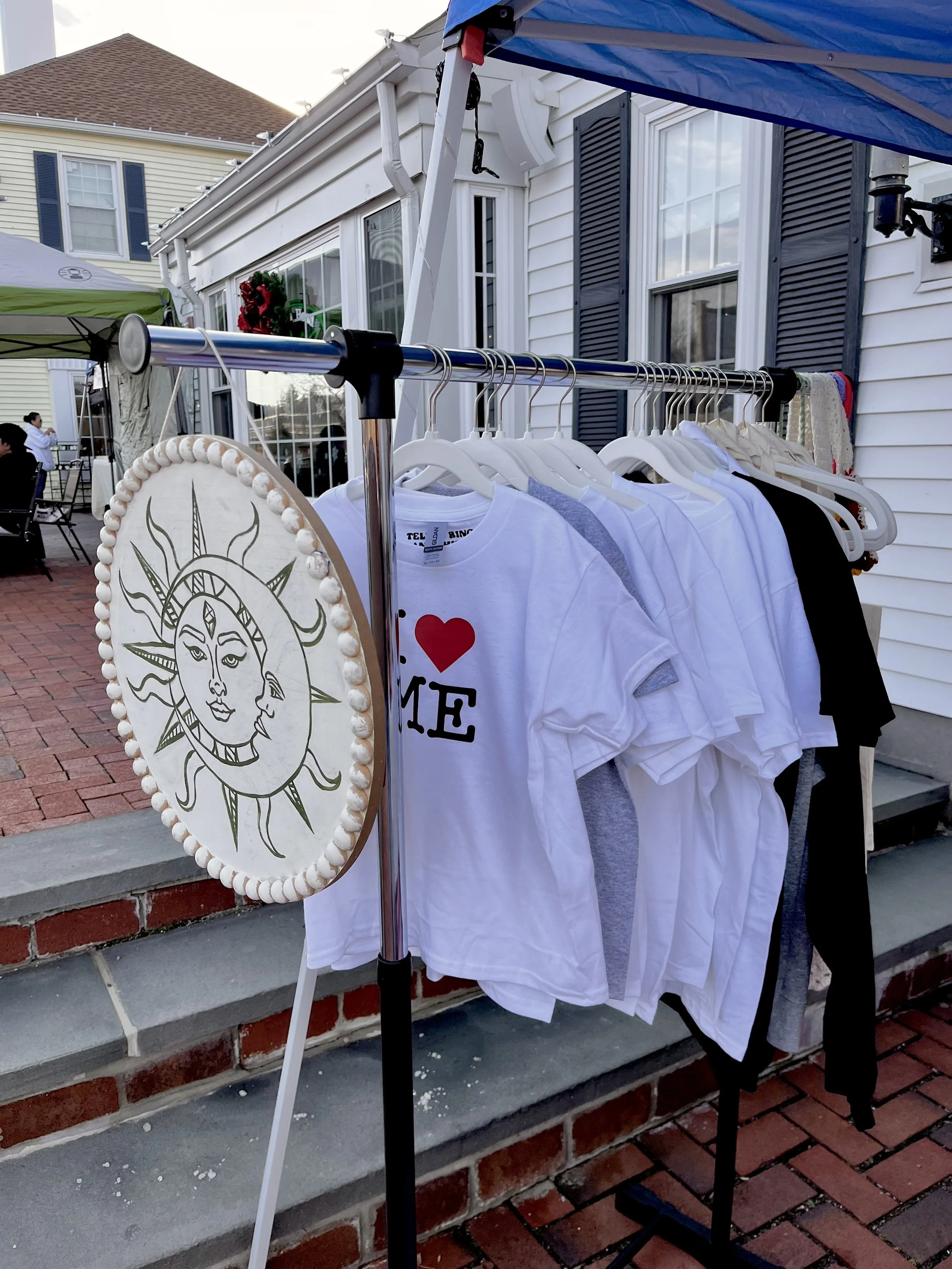 Clothing rack with white and black shirts, including one with red heart and black letters, outdoors in a residential area with houses and a patio.