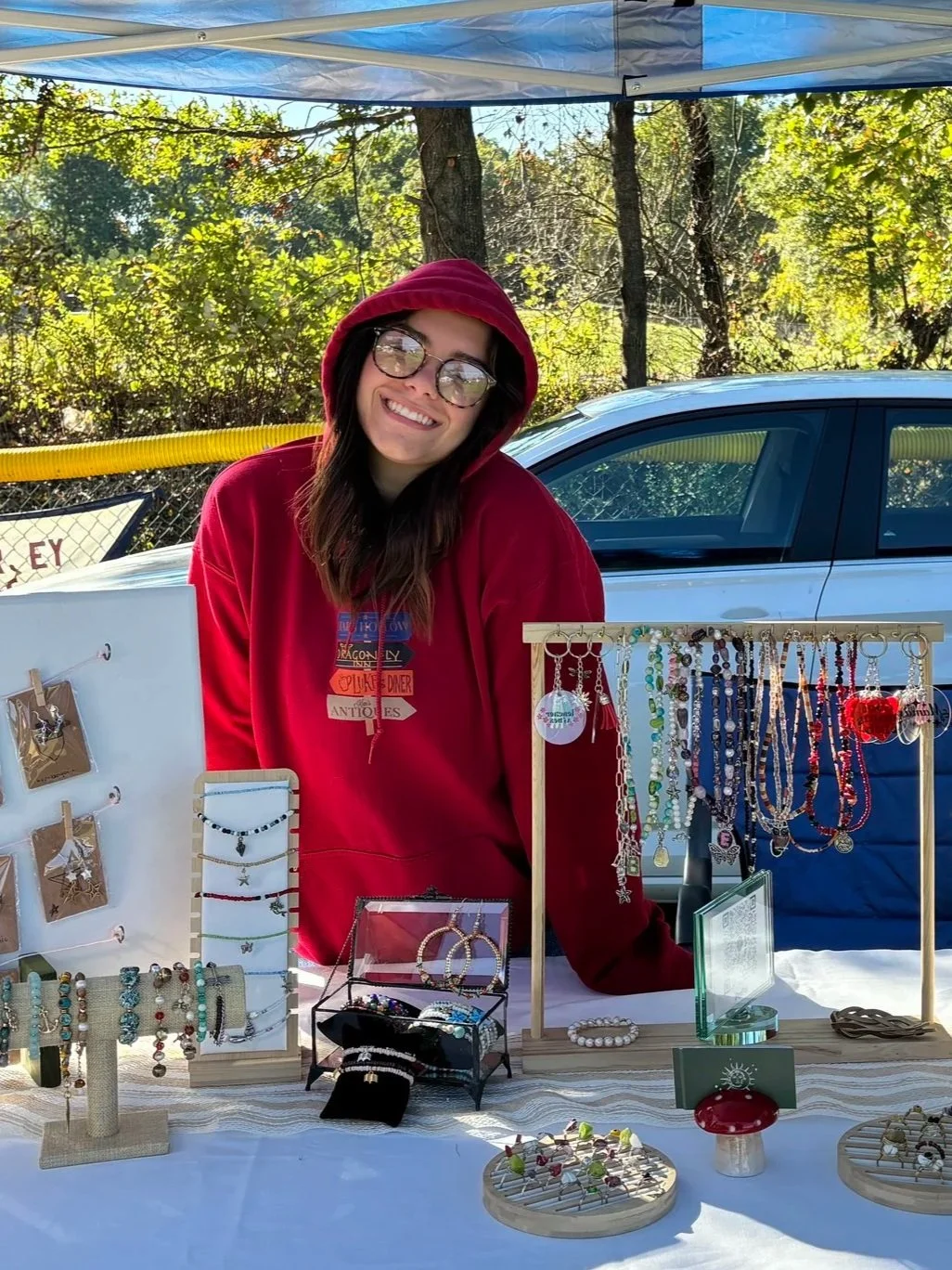 Woman with glasses and a red hoodie standing behind a table with jewelry displayed for sale at an outdoor market, with a car and trees in the background.