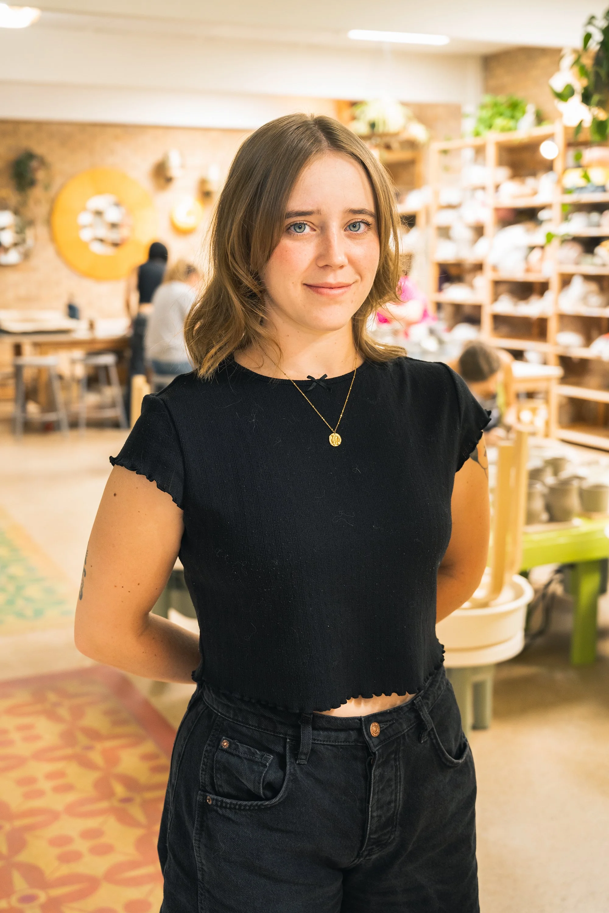 A young woman with shoulder-length brown hair, wearing a black short-sleeve top and black jeans, standing inside a craft or pottery store with shelves of pottery and other items in the background.
