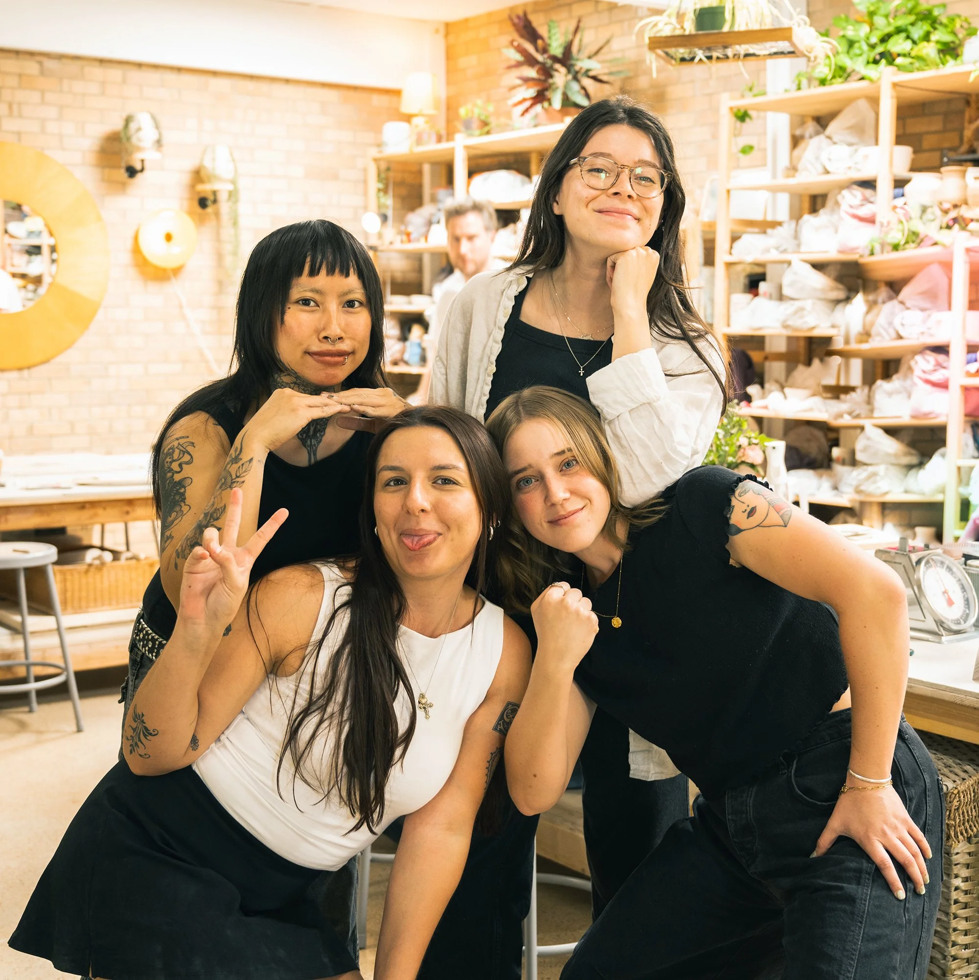 Group of five women smiling and posing for a photo inside a cozy, brightly-lit shop with wooden shelves and brick walls. Some women are making playful gestures, and there are decorative plants and items on the shelves.