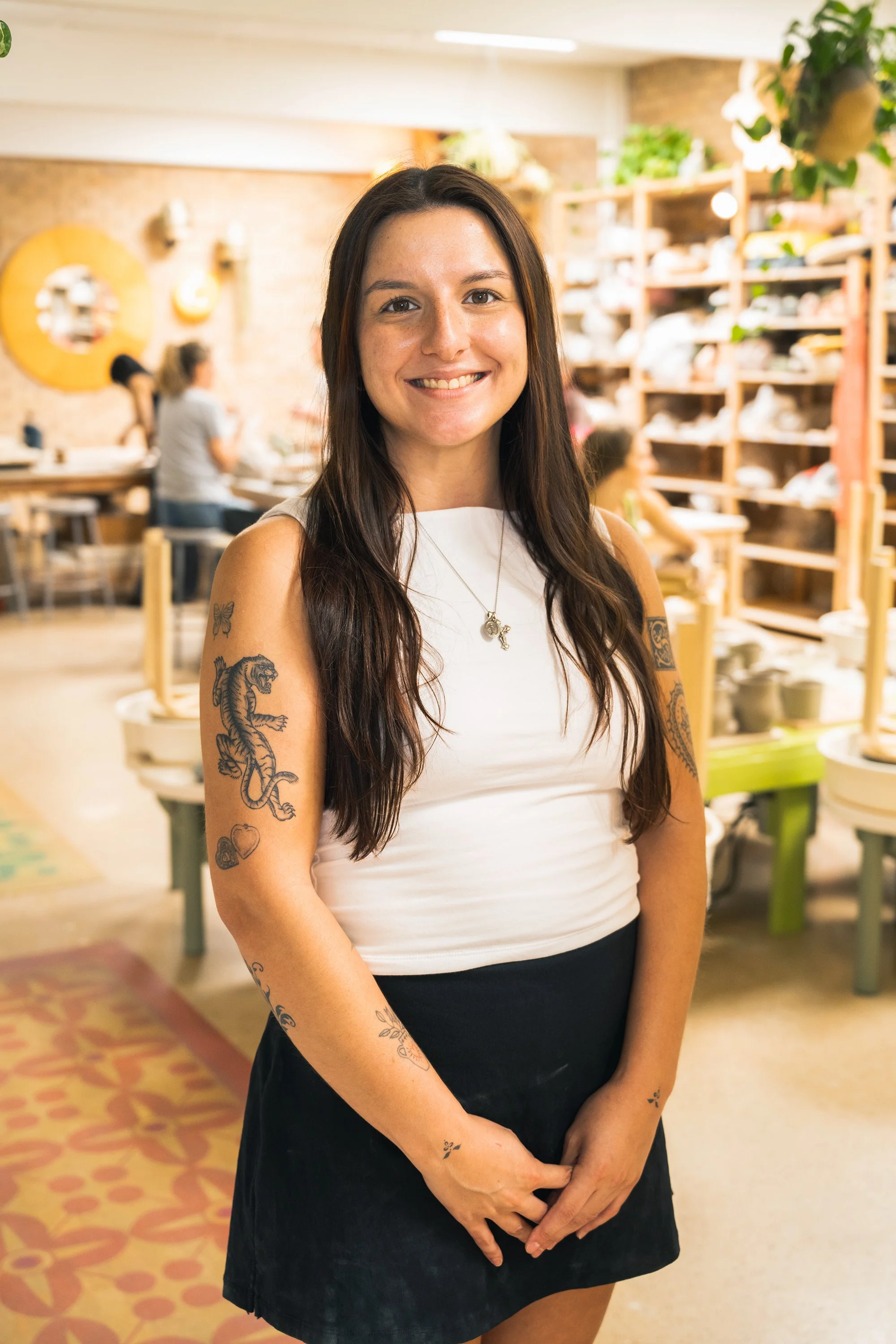 A woman with long dark hair, tattoos on her arms, smiling, standing in a pottery studio with shelves of pottery and other people working in the background.