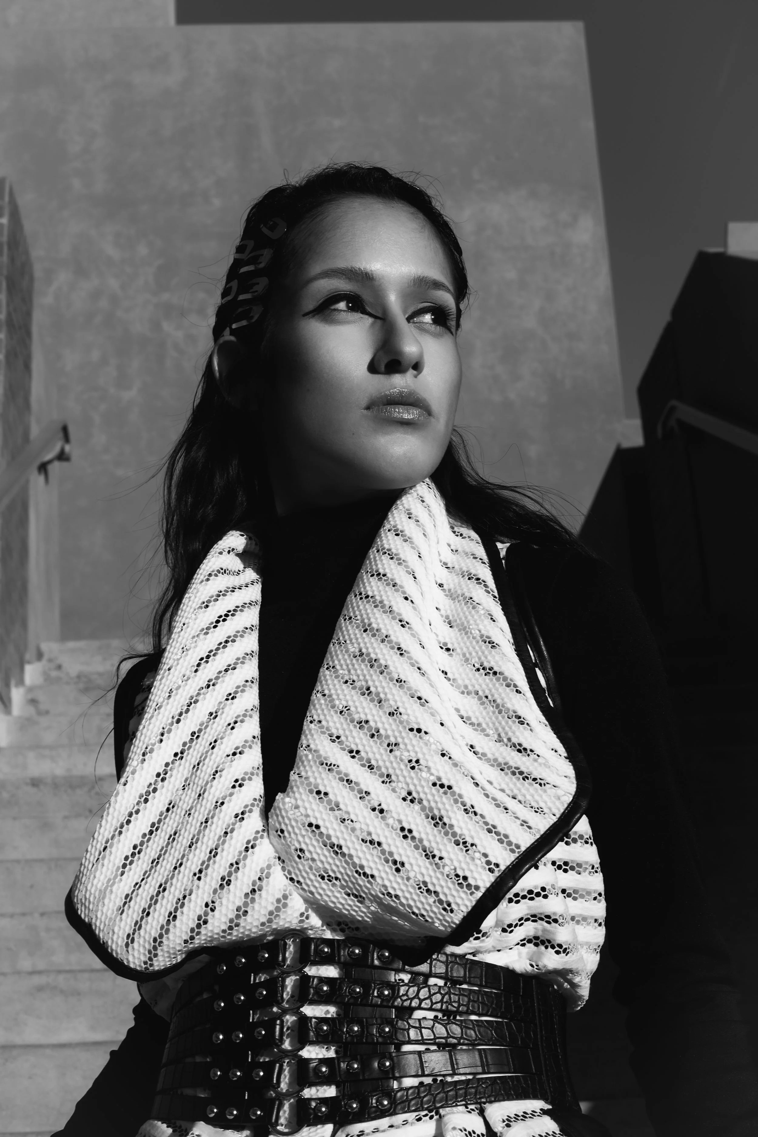 A woman with dark hair and makeup, wearing a patterned white scarf, a black top, and a wide belt, standing outdoors in sunlight with stairs behind her, gazing to the side.