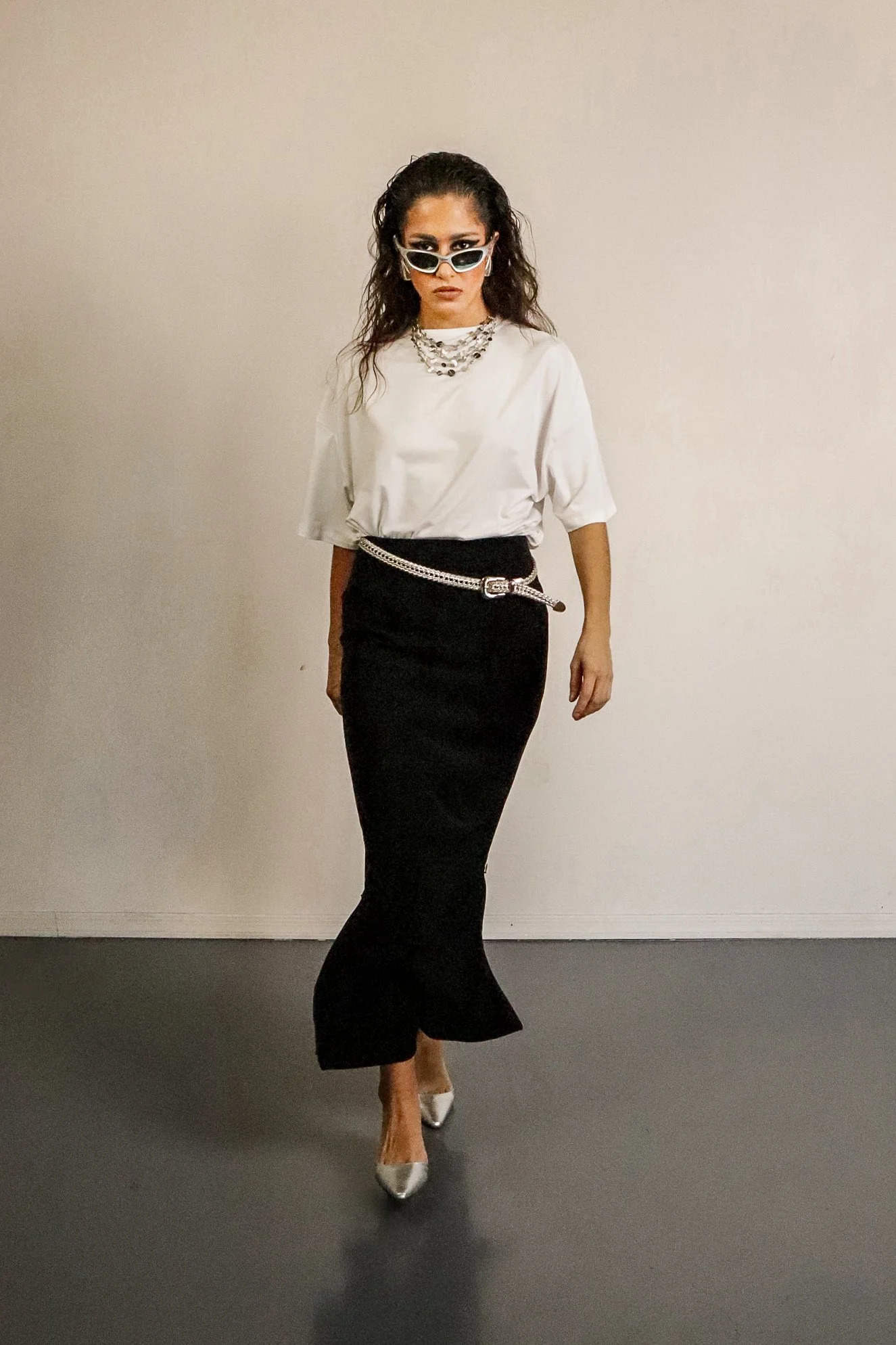 Woman in white T-shirt, black high-waisted skirt with chain belt, silver pointed heels, sporting dark sunglasses, jewelry, and wavy hair, standing against beige wall.