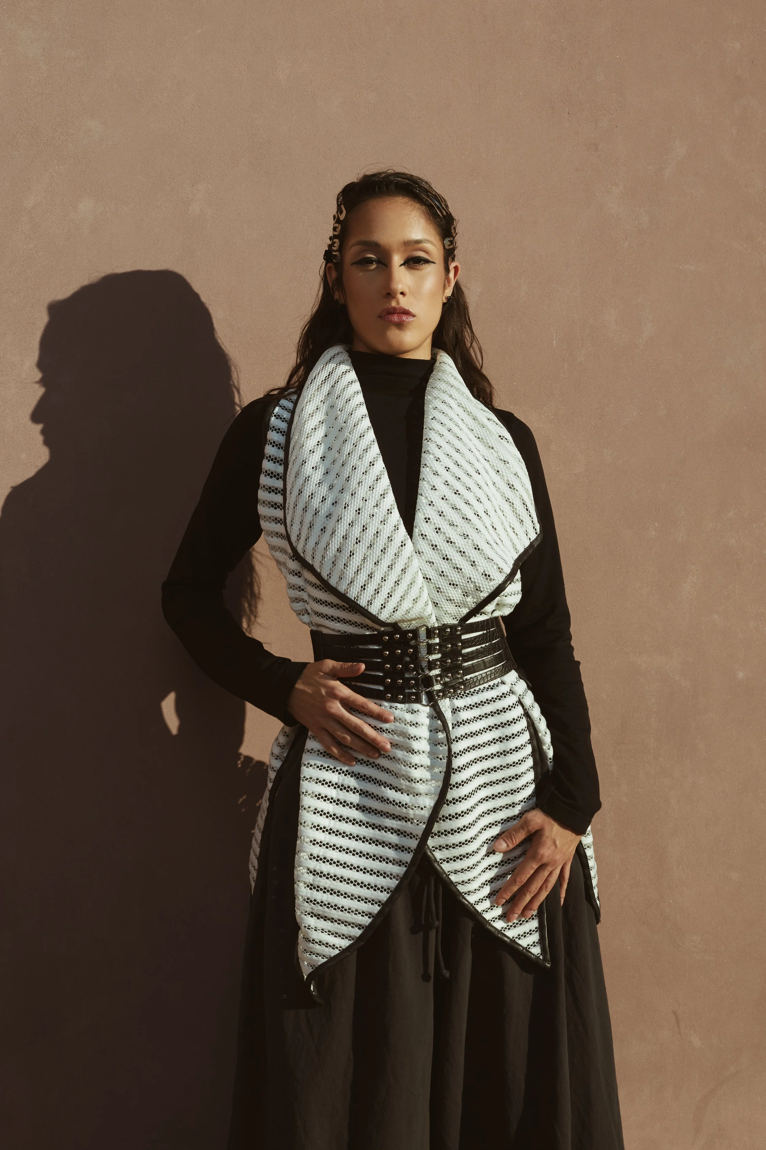 A woman standing against a beige wall, wearing a black and white fashionable outfit with a belt and textured fabric, with her hand on her waist.