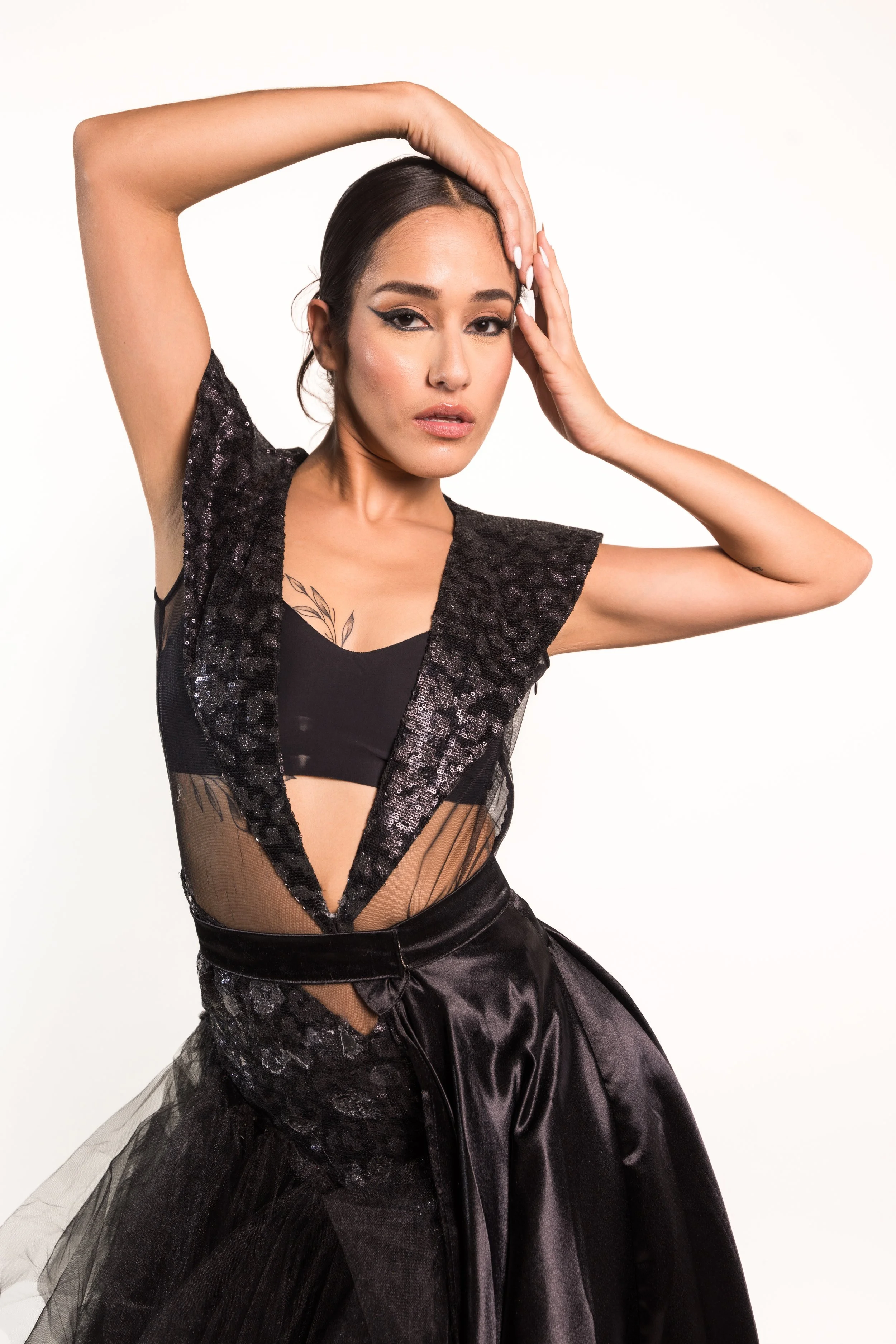 A woman with makeup and dark hair styled back, wearing a black outfit with a deep V-neck and sheer, sequined fabric, striking a pose with one hand on her head and the other touching her face.