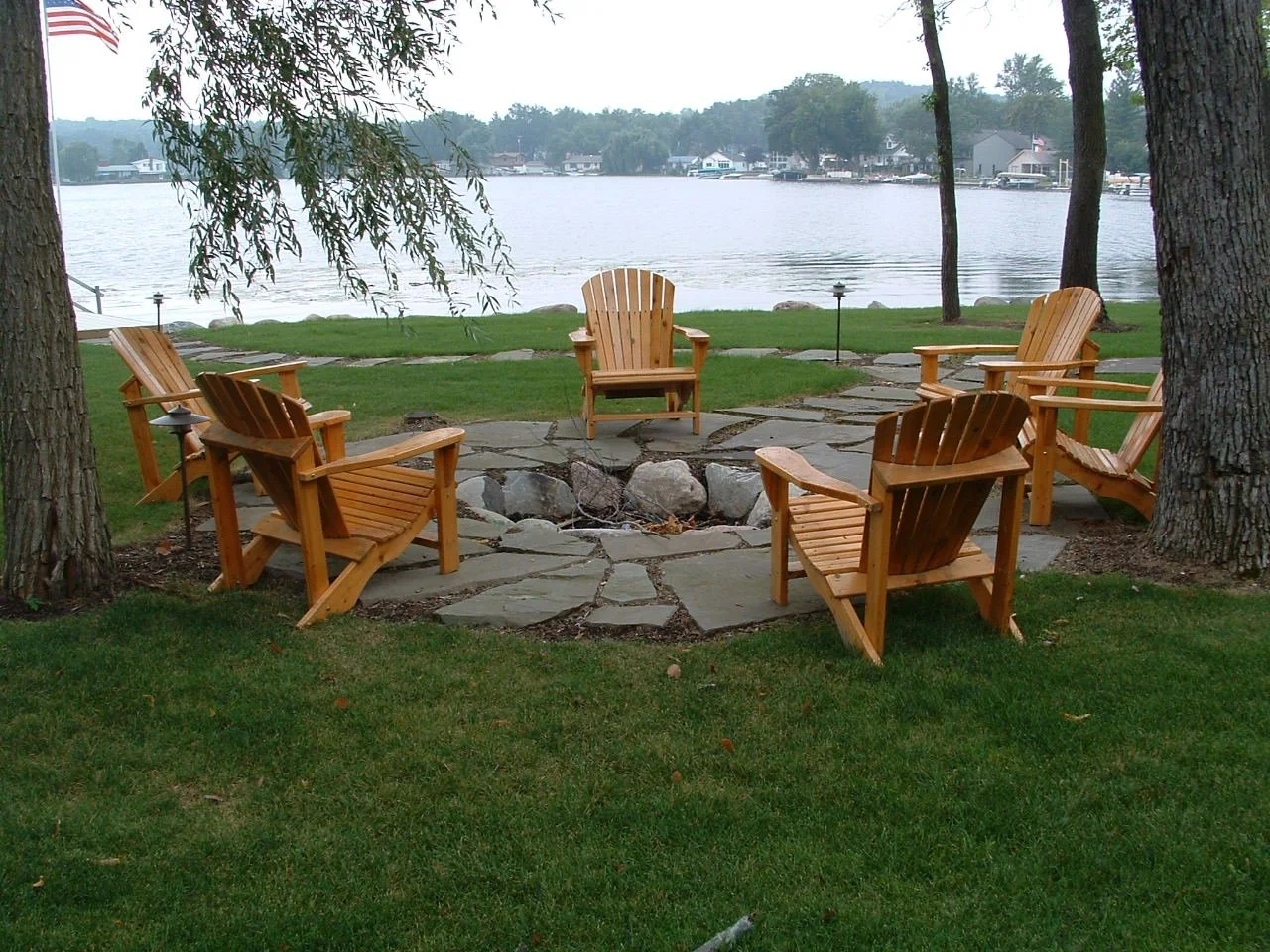 Upcott's Landscaping and Design. Six wooden Adirondack chairs arranged in a circle around a stone fire pit on a grassy lakeside lawn with trees and water in the background.