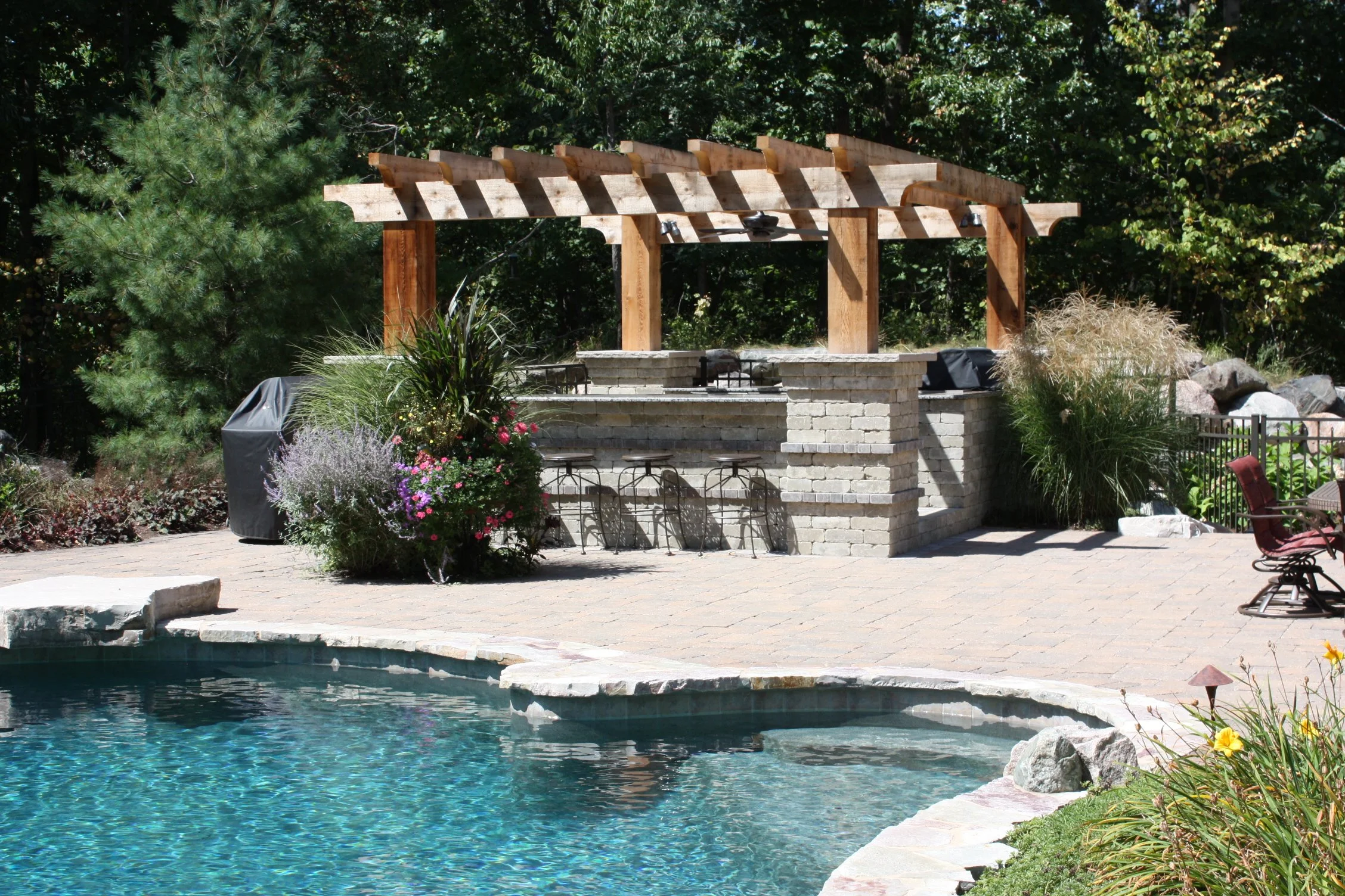 Upcott's Landscaping and Design. Backyard swimming pool with a stone and brick bar area, wooden pergola, plants, trees, and outdoor seating.