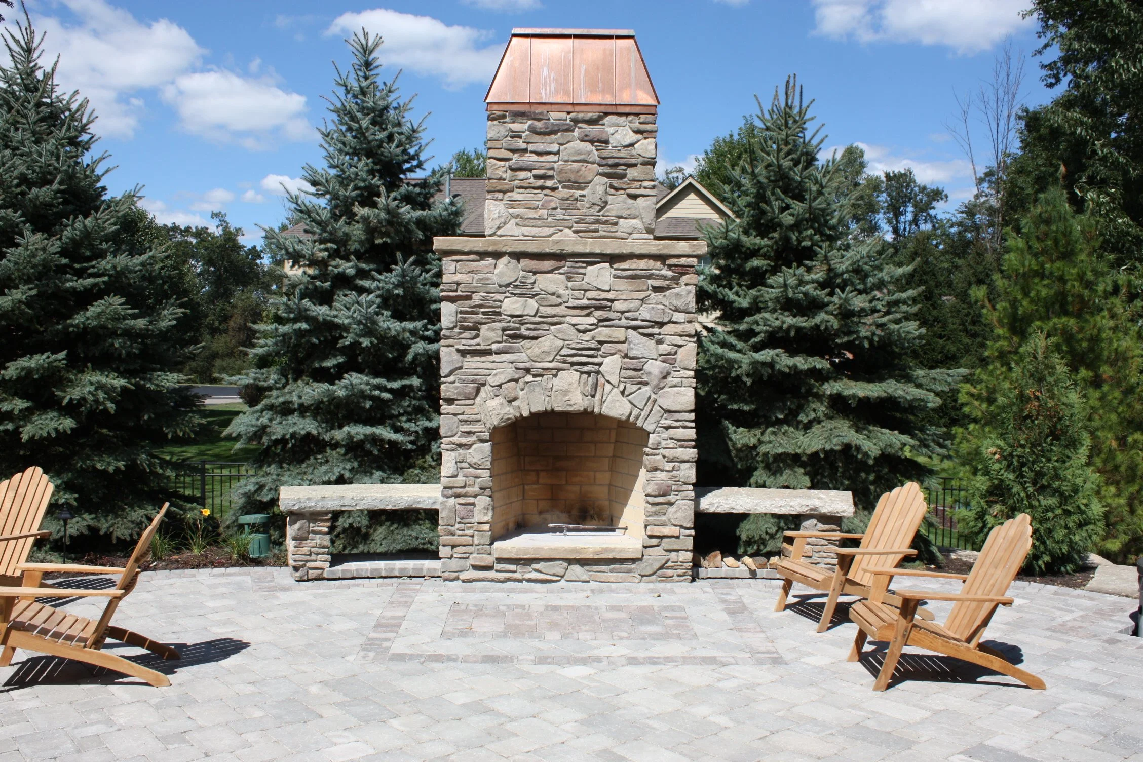 Upcott's Landscaping and Design. A stone outdoor fireplace with a copper roof, surrounded by pine trees and four wooden chairs on a paved patio.