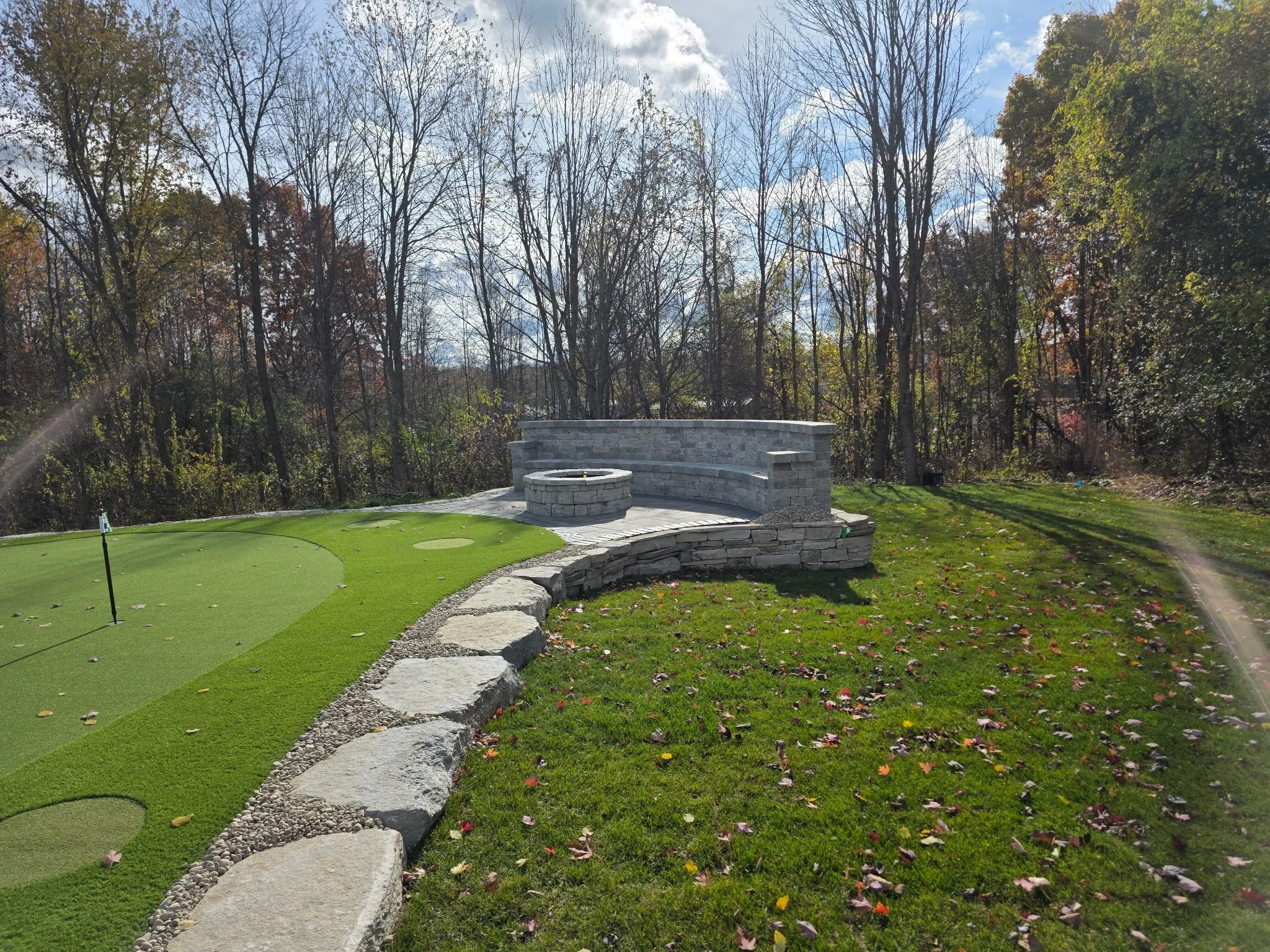 A backyard putting green with a curved stone border and a stone fire pit area, surrounded by leafless trees and a partly cloudy sky.