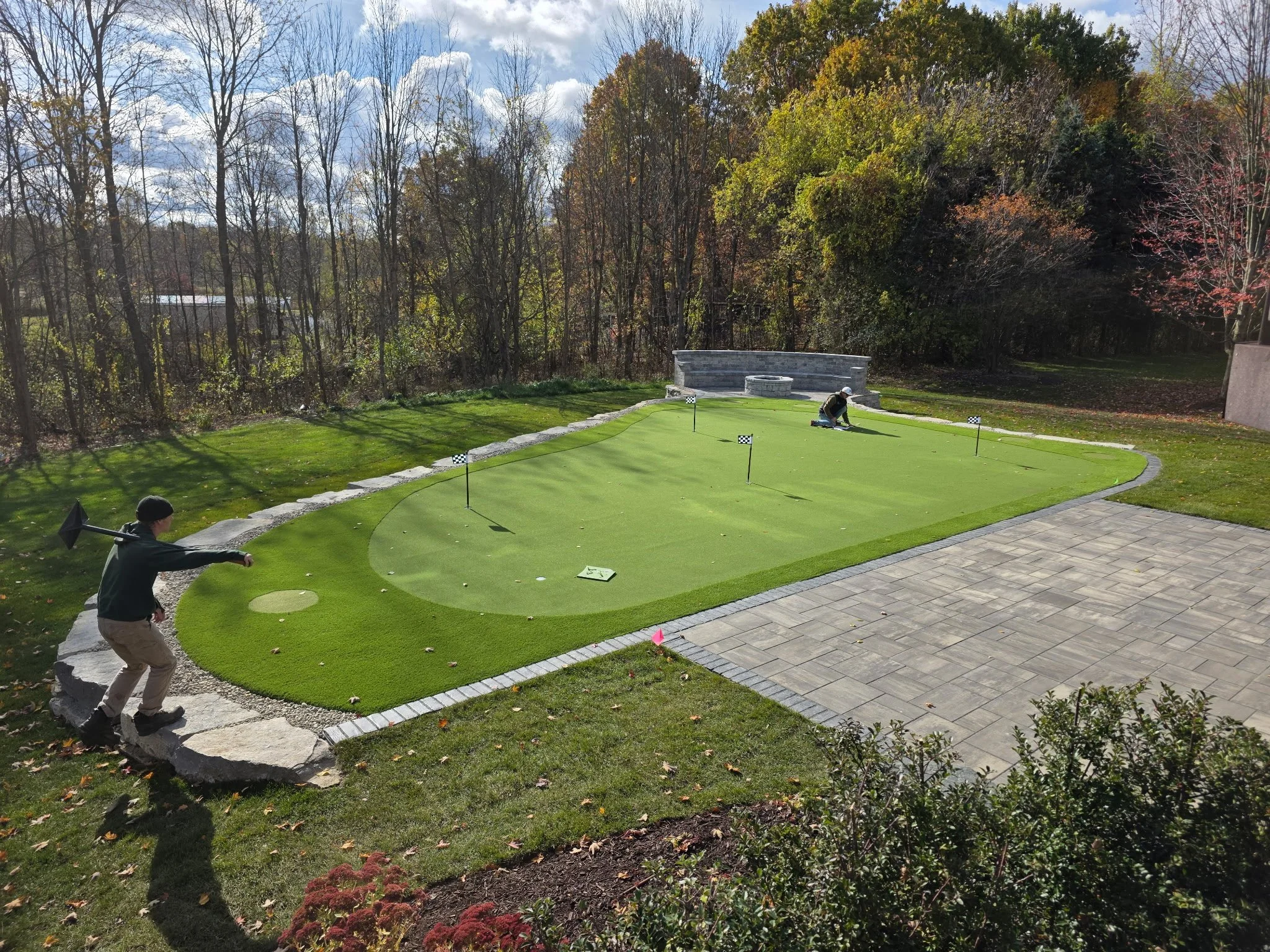 People maintaining a miniature golf putting green outdoors on a sunny day, with trees in the background and a stone patio area.