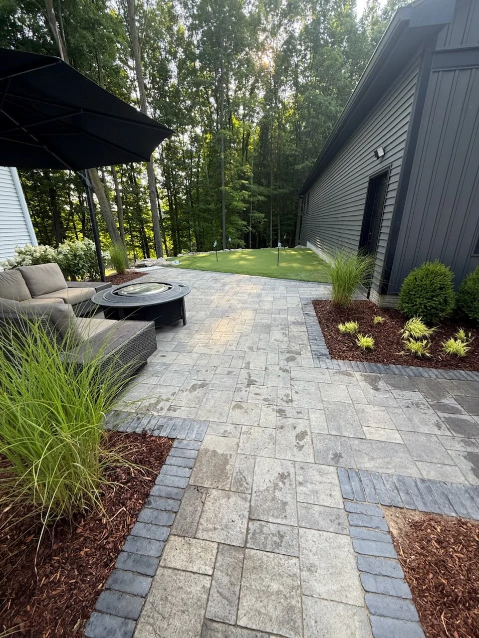 Backyard patio with stone paving, a black umbrella, outdoor sofa and fire pit, green plants and shrubs, and a small putting green with flags, surrounded by trees.