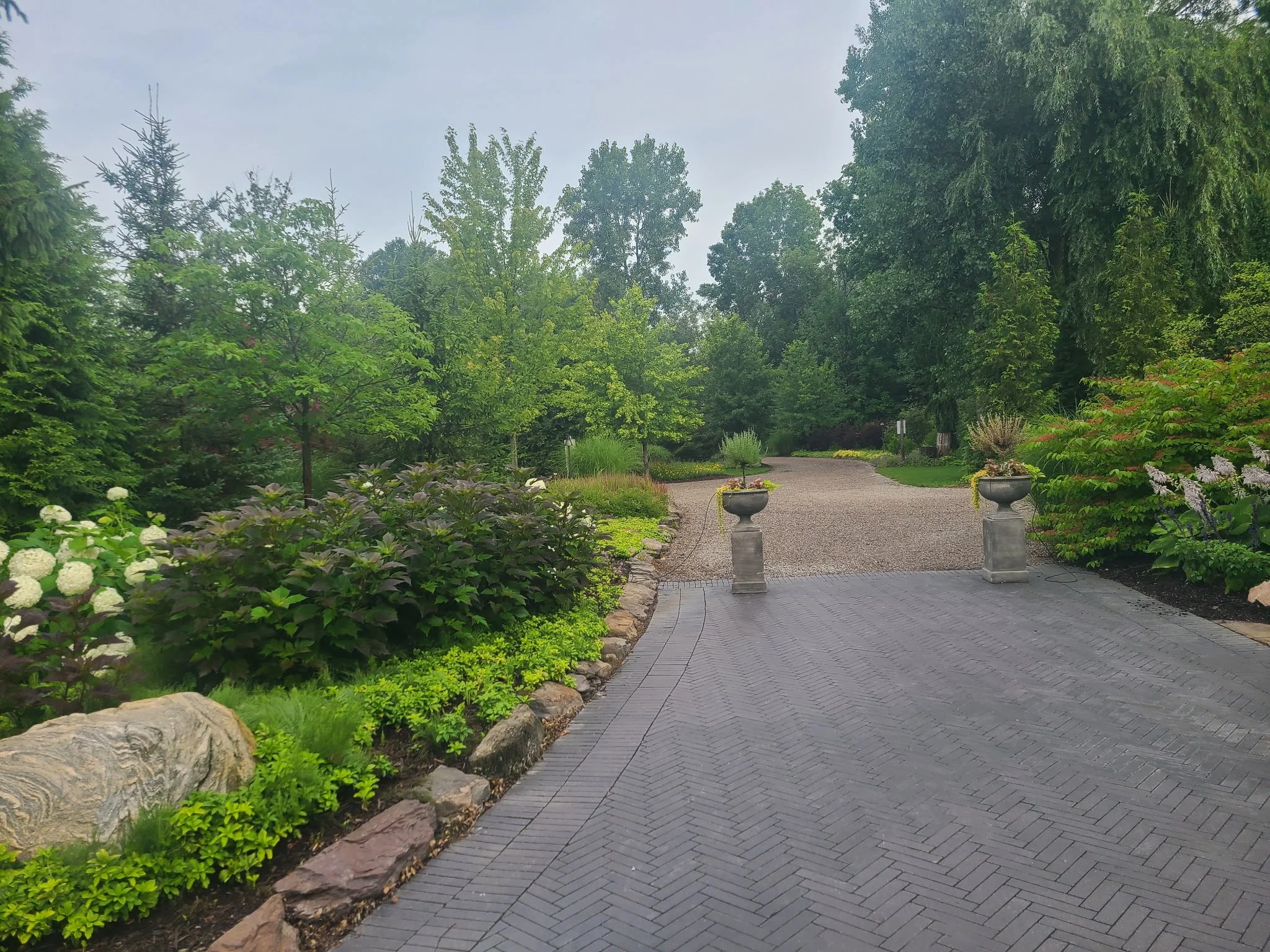 A landscaped garden pathway with stone paving, decorative stone planters, and lush green trees and bushes surrounding the area.