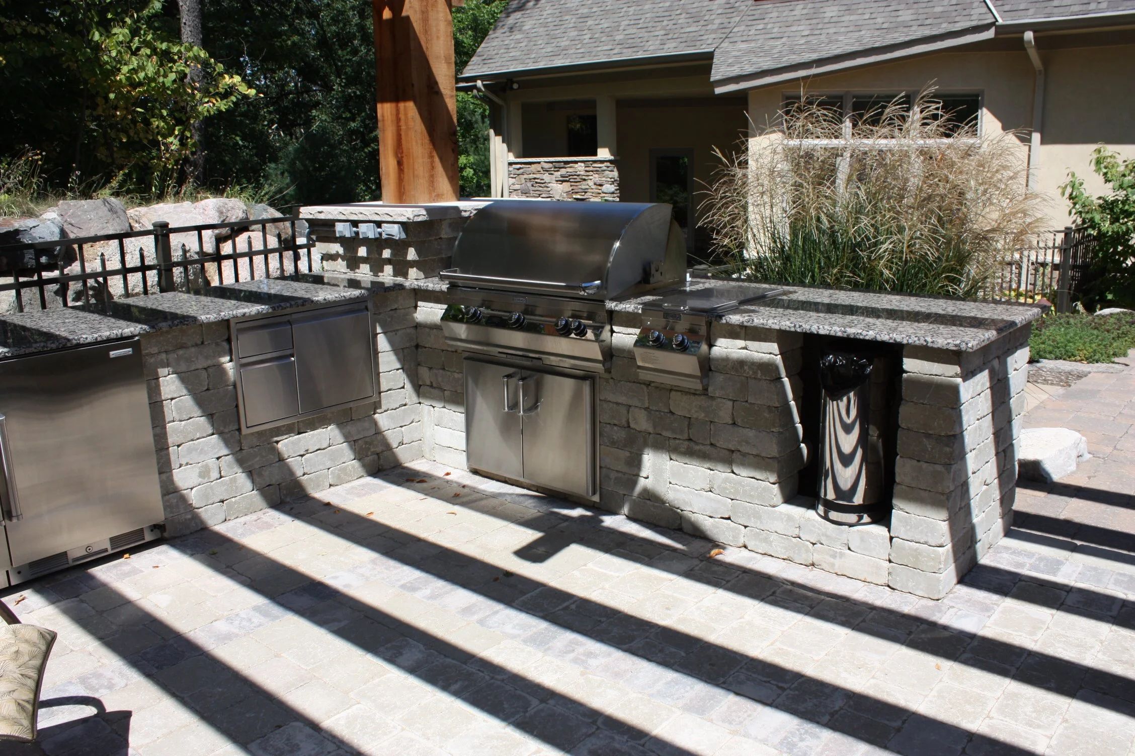 Upcott's Landscaping and Design. Outdoor kitchen with stainless steel appliances, brick counter, granite countertop, and surrounding plants.