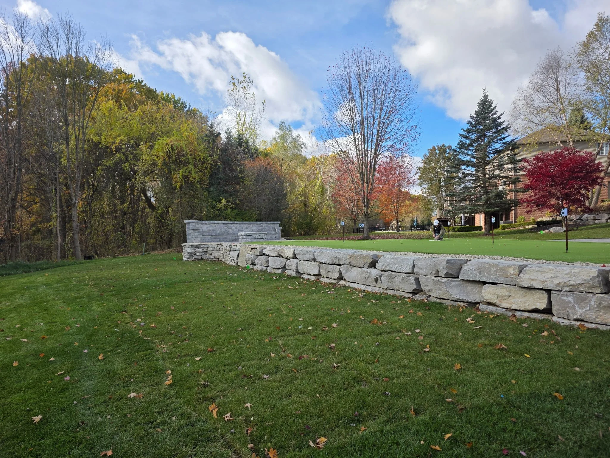 View of a golf course with a stone wall, trees with fall foliage, a person playing golf, and residential buildings in the background under a partly cloudy sky.