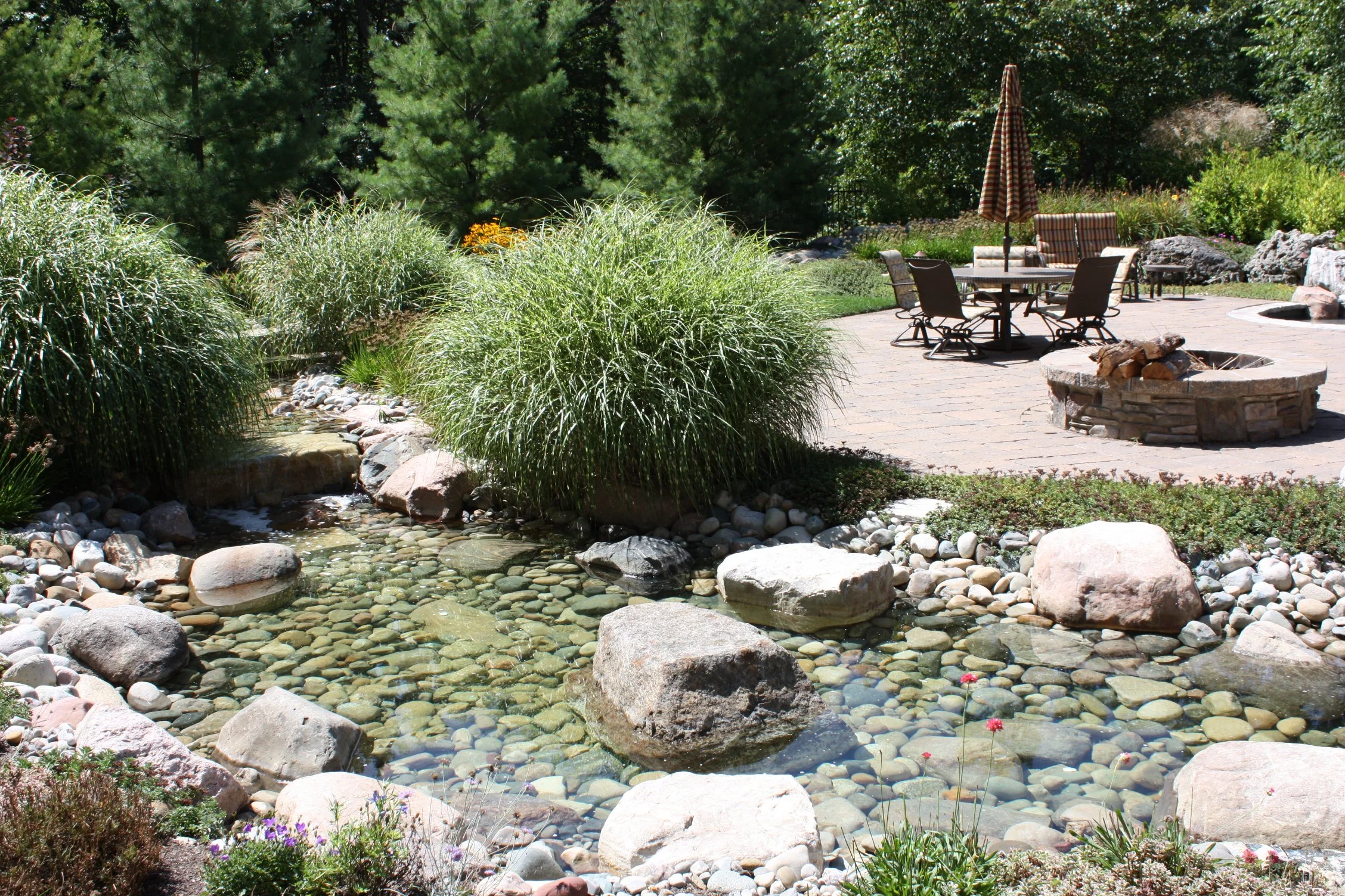 Upcott's Landscaping and Design. Backyard with a pond, large rocks, grassy bushes, and a stone fire pit on a brick patio with outdoor furniture and a striped umbrella.