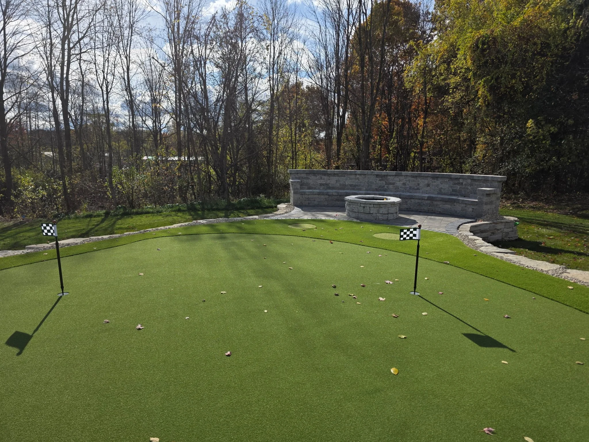 Miniature golf putting green with two checkered flags, a stone wall backdrop, surrounded by fallen leaves and wooded area with trees in autumn.