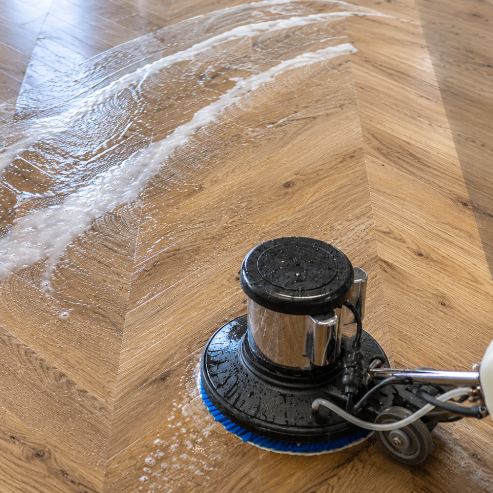 Close-up of a floor cleaning machine scrubbing a wooden floor with soap and water.