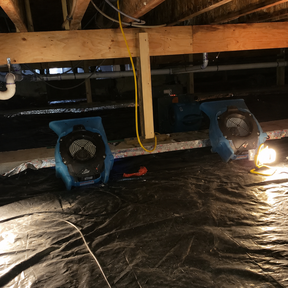 Construction site with two large blue industrial fans, a bright work light, wooden beams, pipes, and wiring, on a black plastic ground covering.