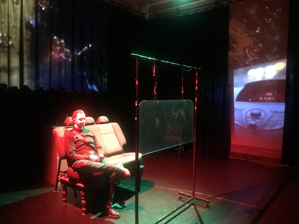  A wider image of the space with David sitting on the car seat, there is a projection of a road on the windscreen. 
